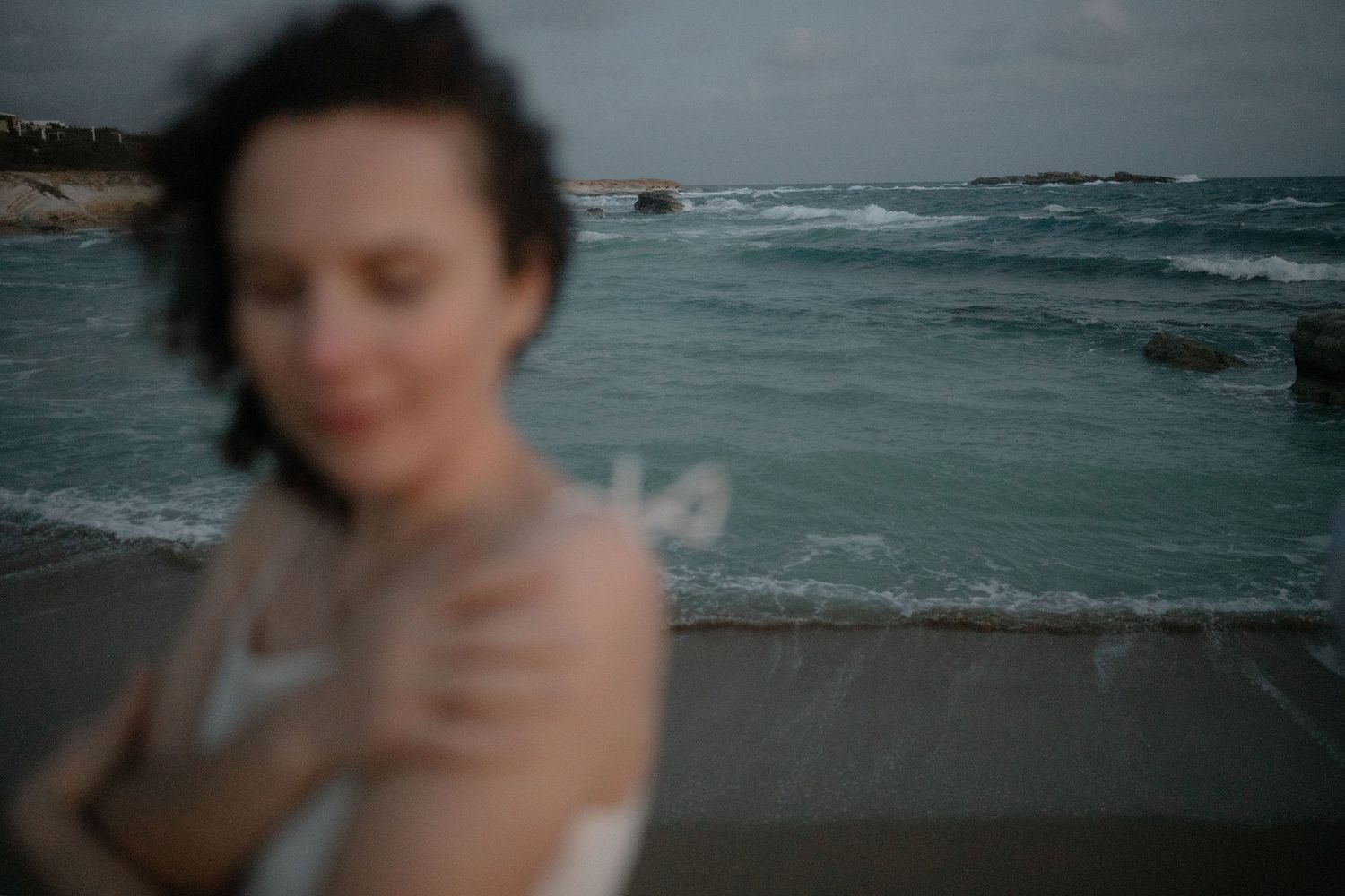 Blurred woman in white dress stands near rocky ocean shore with turquoise waves under overcast sky.