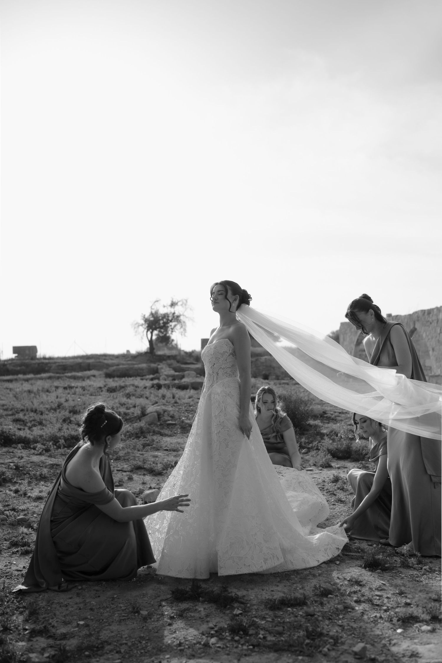 Black and white photo of a bride in a lace gown with bridesmaids adjusting her veil outdoors.