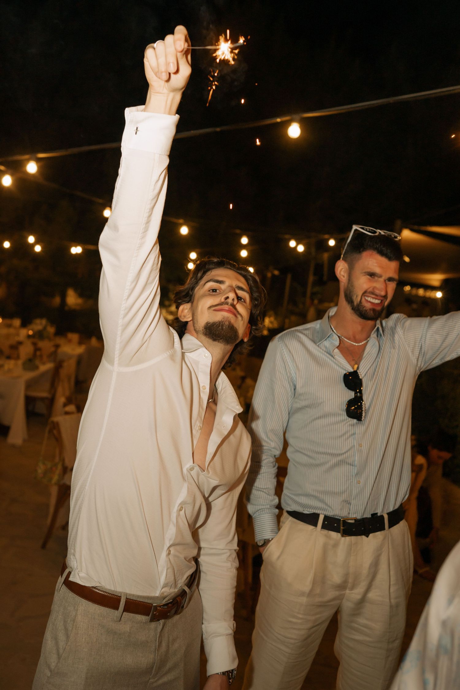 Two men celebrating at an outdoor evening wedding reception, one holding a sparkler aloft under string lights.