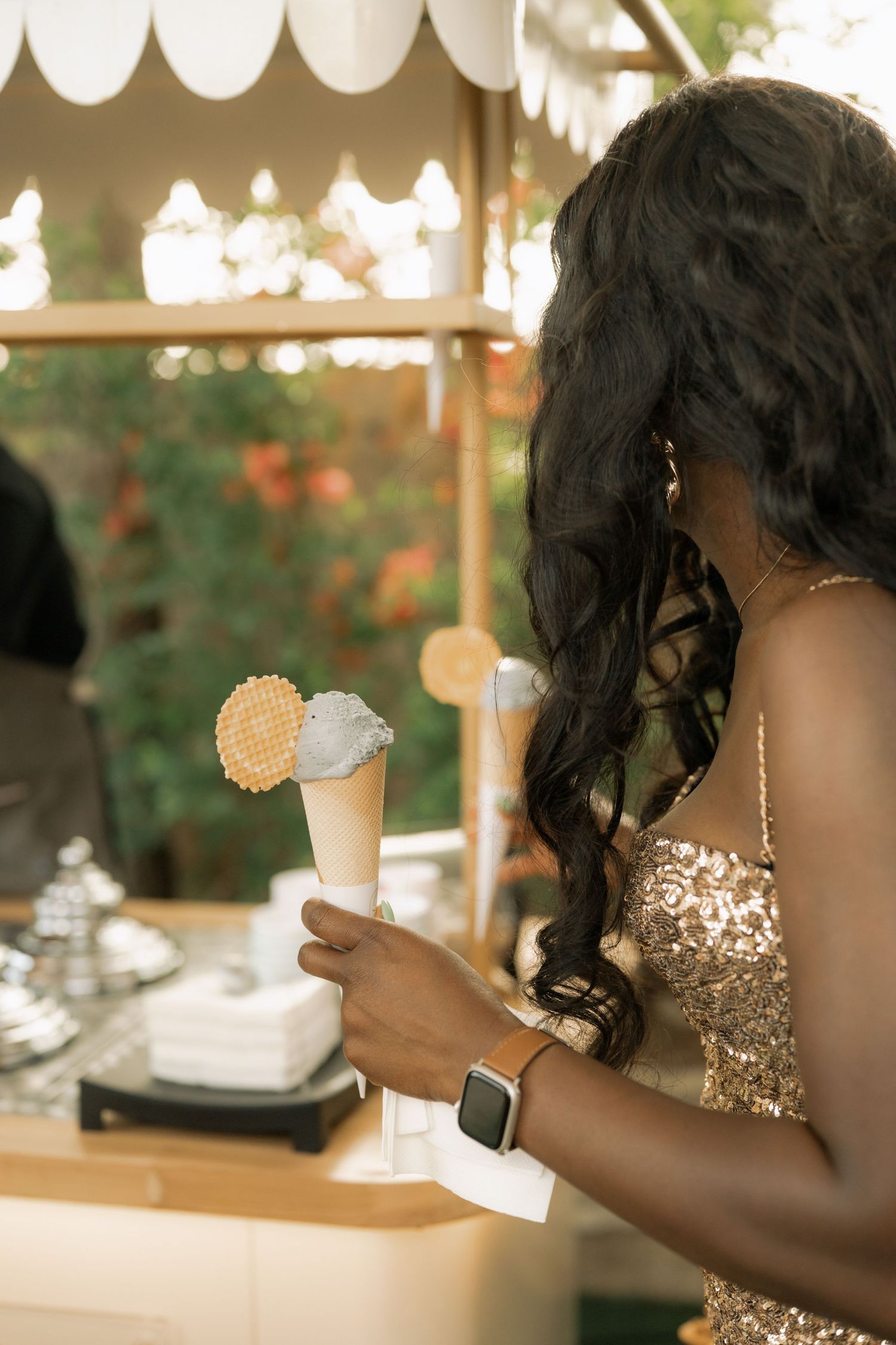 Woman in gold sequin dress holding ice cream cone at elegant outdoor dessert stand.