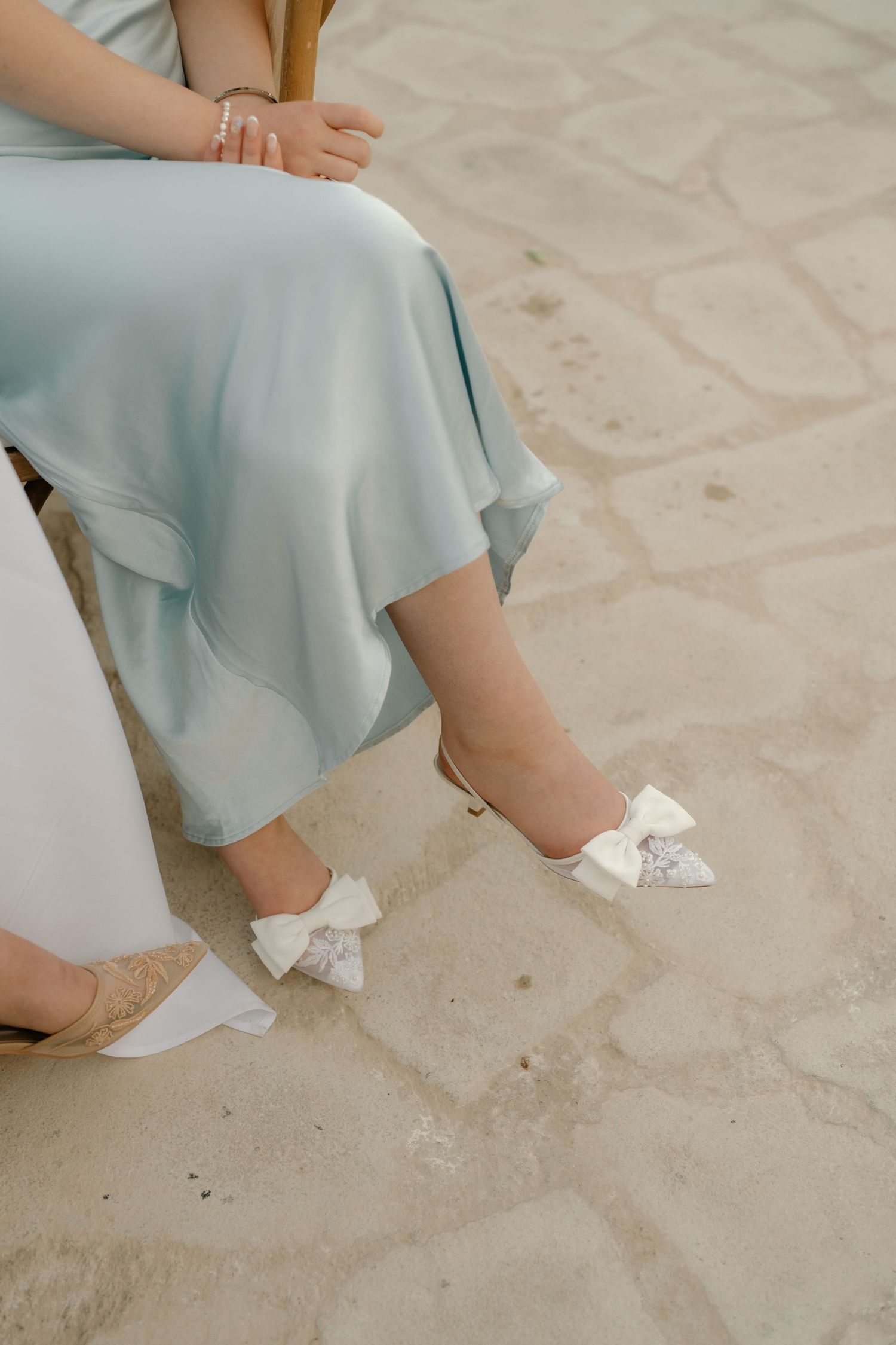 Woman in light blue satin dress wearing white bow heels seated on stone patio.