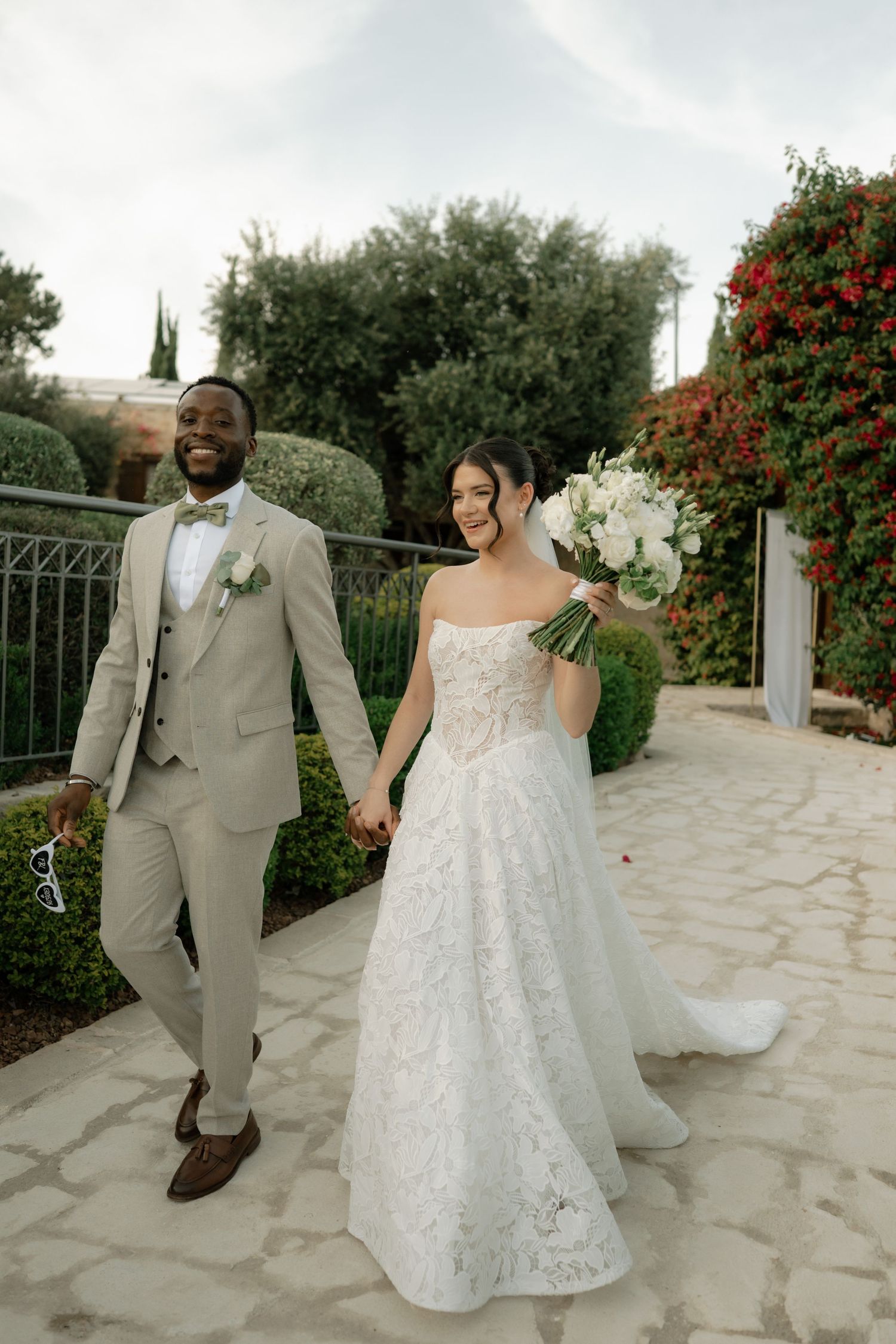 Bride and groom walking hand in hand outdoors, groom in tan suit, bride in white lace gown holding white bouquet.
