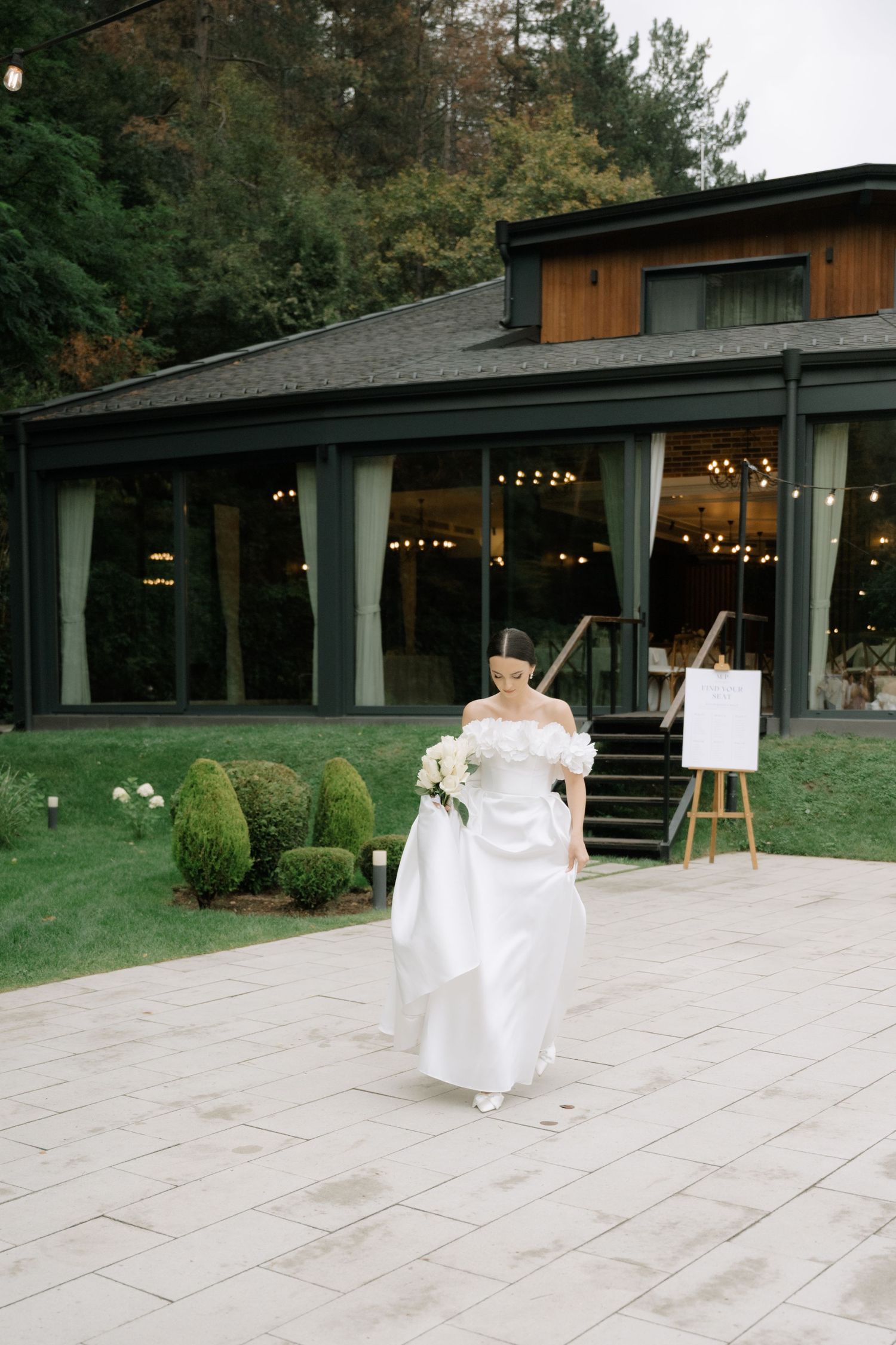 A bride in a white off-shoulder wedding gown walks on a stone patio outside a modern venue with string lights and topiary bushes.