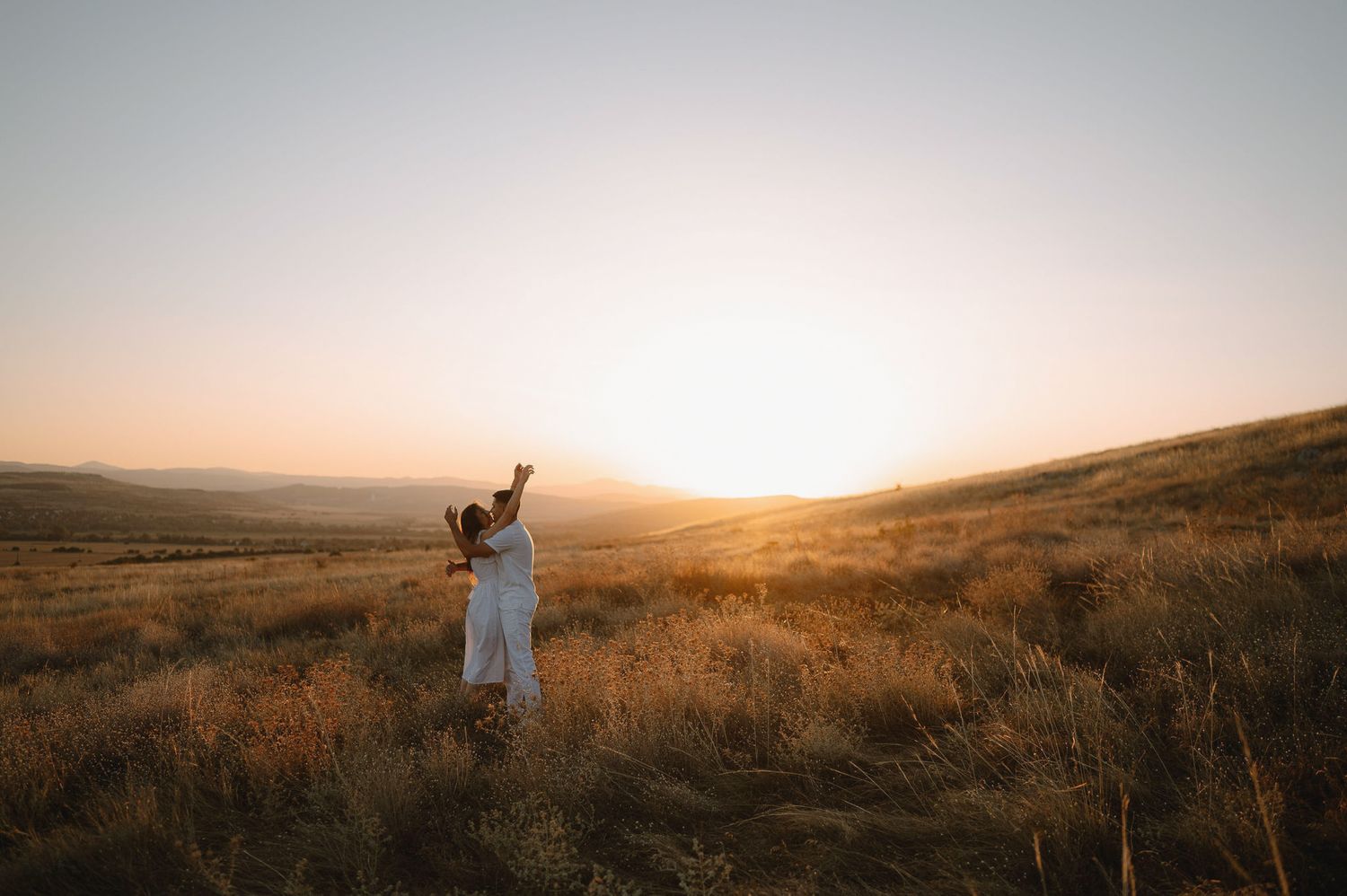 Couple embraces in golden field at sunset with dramatic mountain landscape in background.