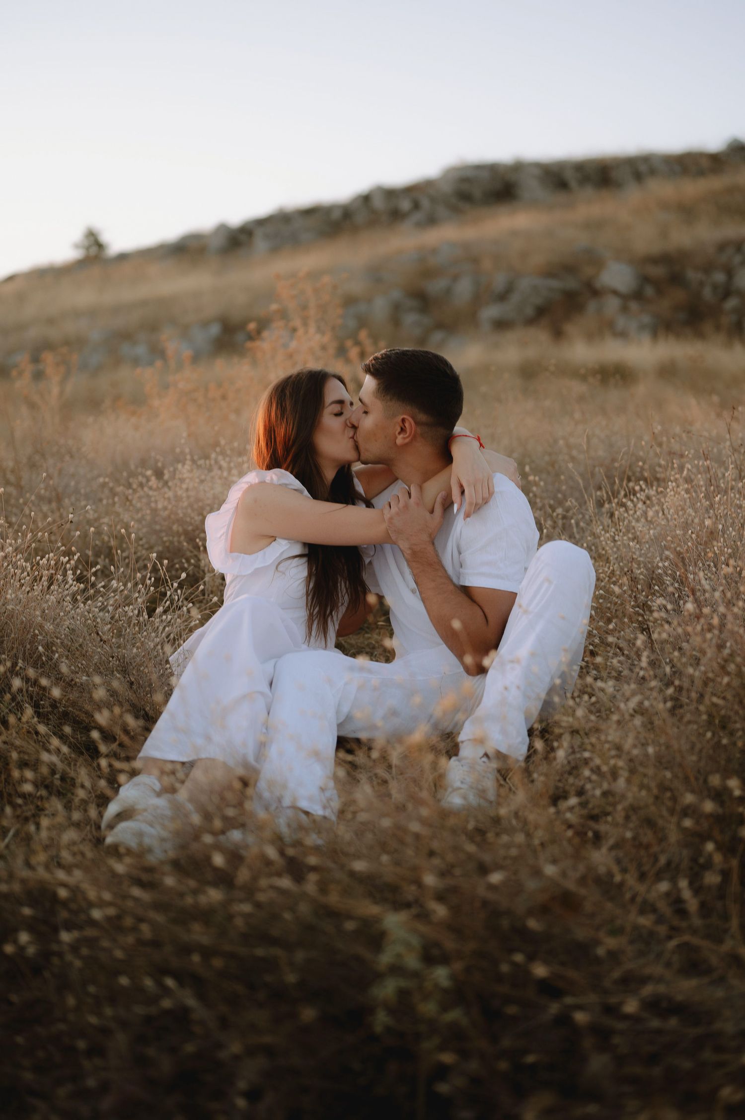 A romantic couple in white outfits share an intimate moment while sitting in a golden field at sunset.