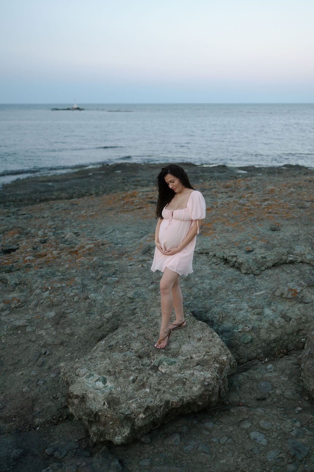 A light pink dress flows in the coastal breeze as someone stands on rocky shoreline at dusk with calm ocean waters behind.