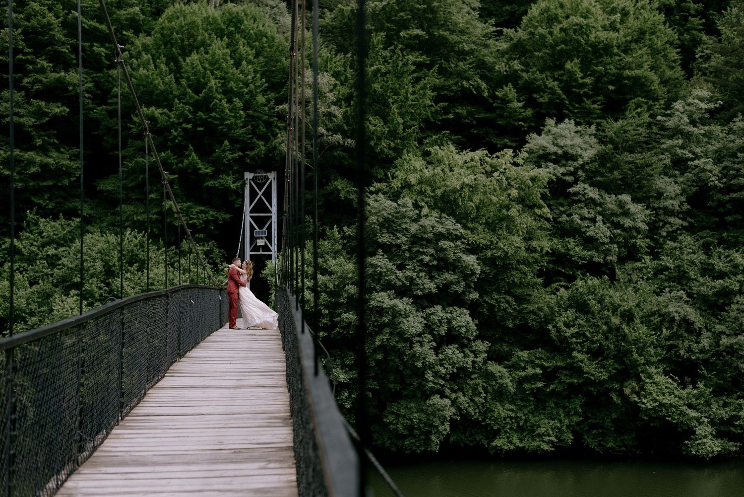 A couple in wedding attire walks across a scenic suspension footbridge surrounded by lush green forest.