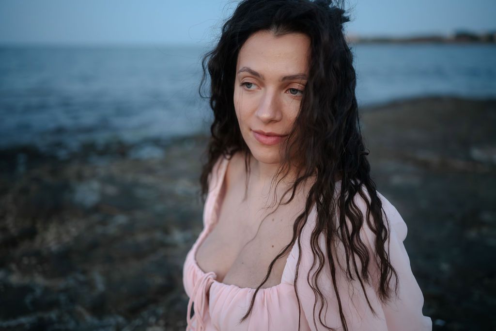 Portrait of someone with dark wavy hair wearing pink against a moody coastal background at dusk.