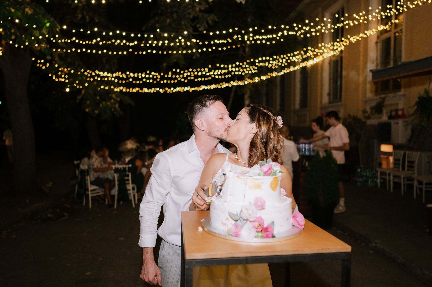A couple shares a kiss next to their pink floral wedding cake under twinkling string lights at an evening reception.