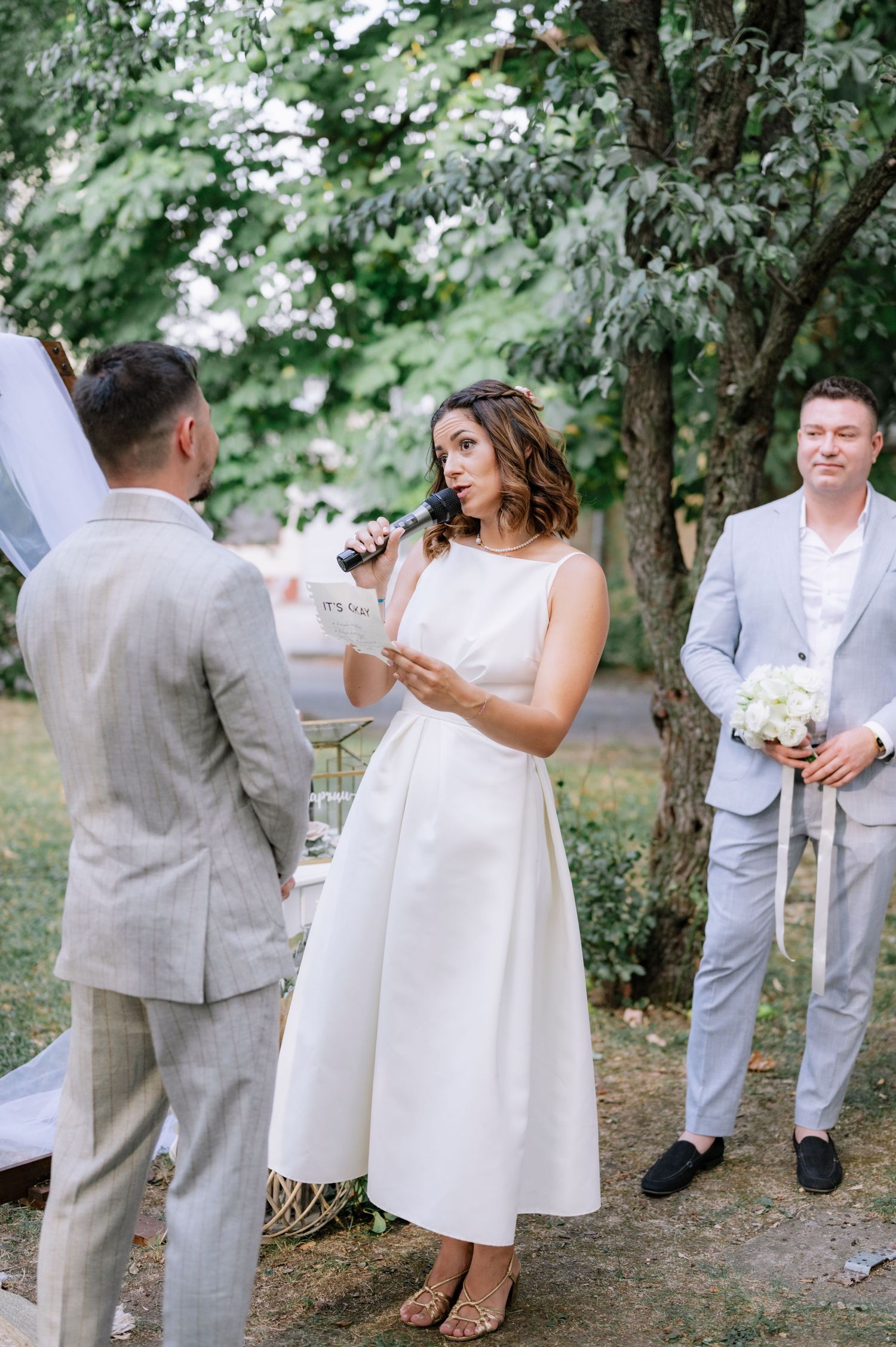 A wedding ceremony in a garden setting with people in light colored formal attire exchanging vows among trees.
