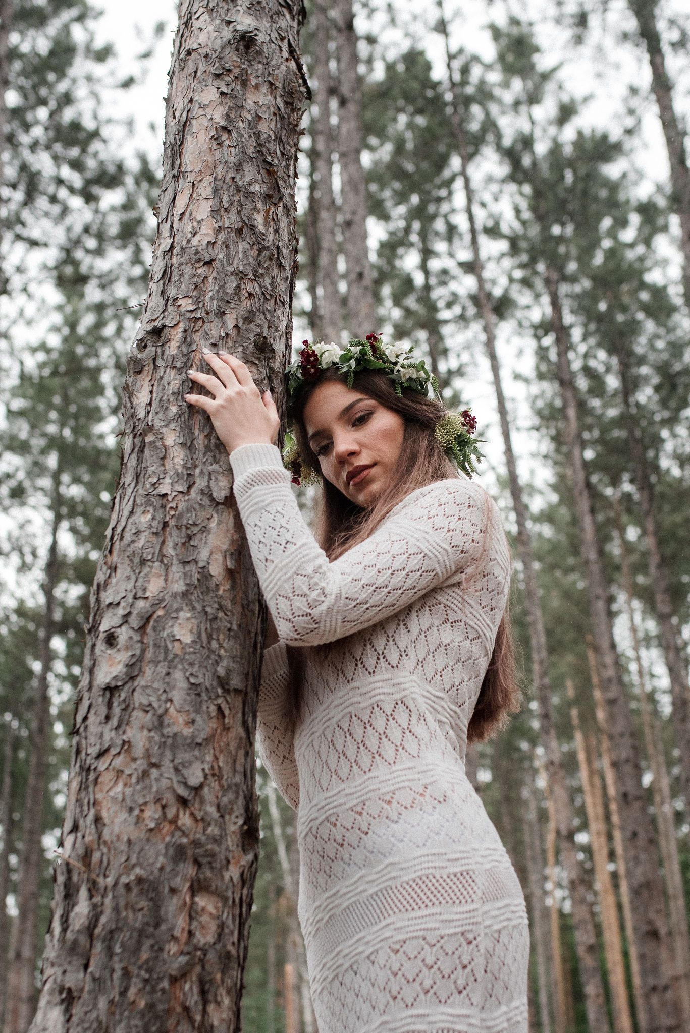 A woman in a white lace dress poses gracefully against pine trees in a forest setting.