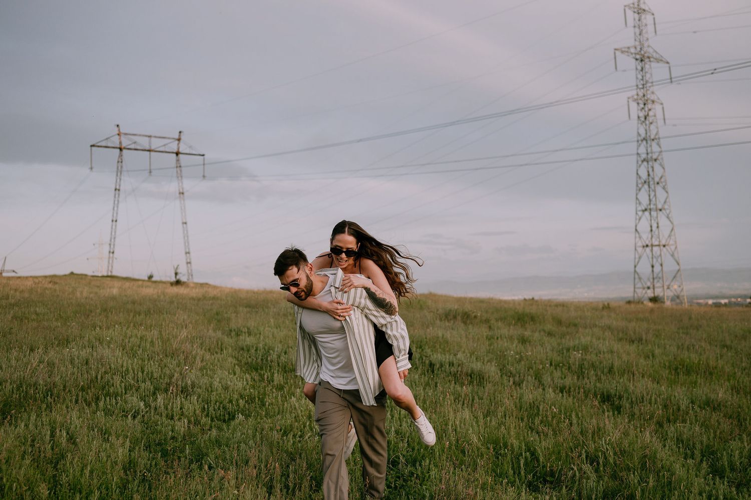 A playful couple enjoys a piggyback ride in a grassy field beneath power lines and transmission towers at dusk.