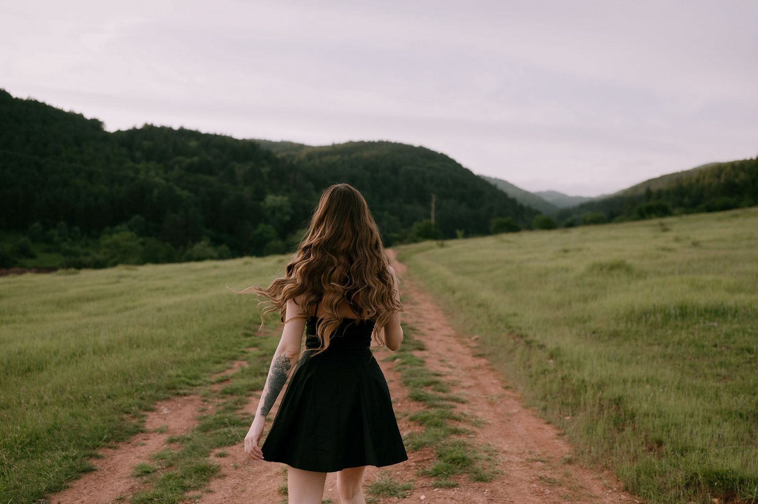 Person in a black dress stands on a dirt path between green hills under an overcast sky.