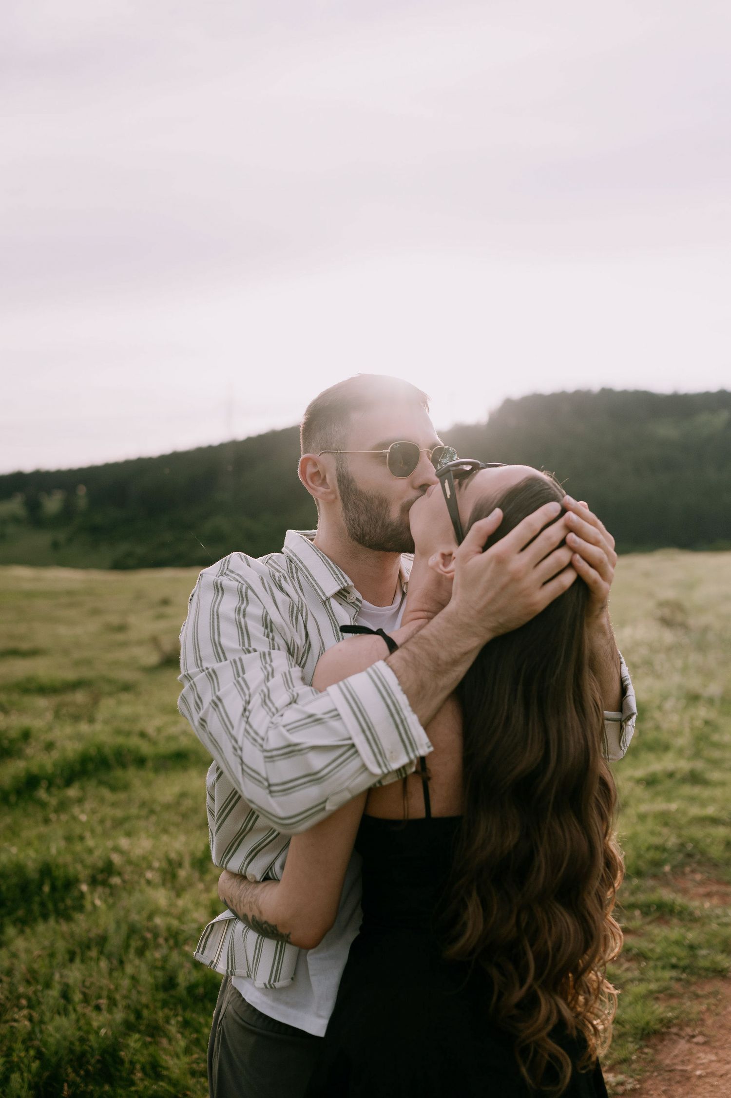A romantic couple embraces and kisses in a serene field with mountains and fog in the background.