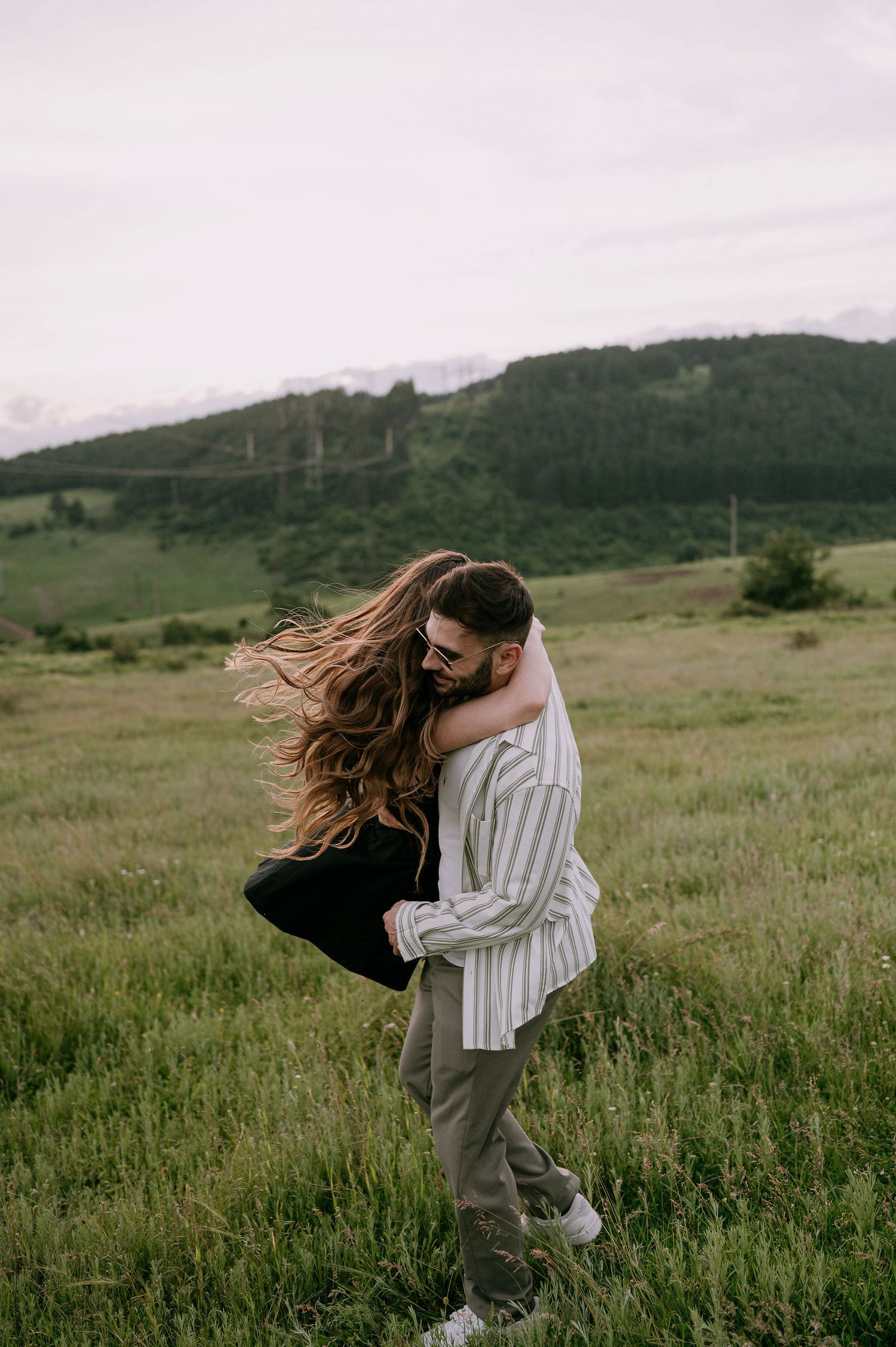 A couple embraces in a windswept meadow with rolling green hills in the background during golden hour.