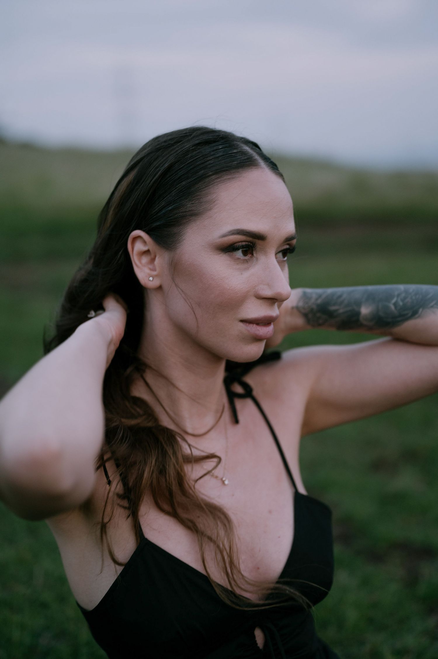 Woman in black top poses outdoors at dusk in a grassy field with soft natural lighting.