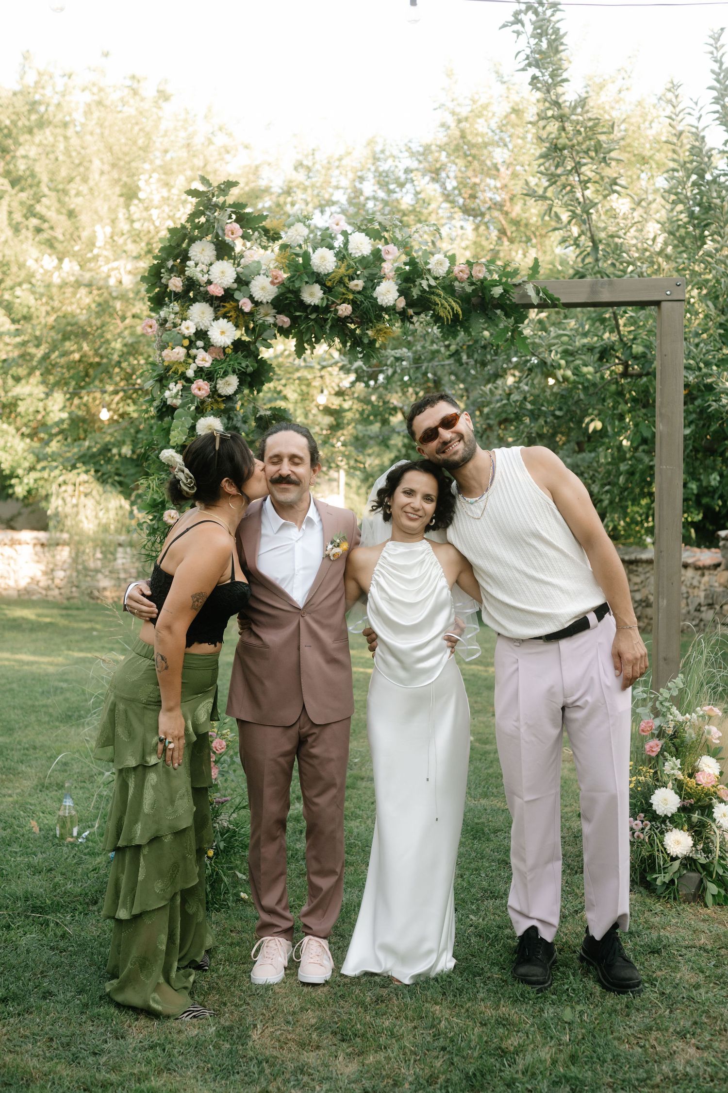 A wedding party poses together outdoors in brown and white attire against a backdrop of blooming roses and greenery.