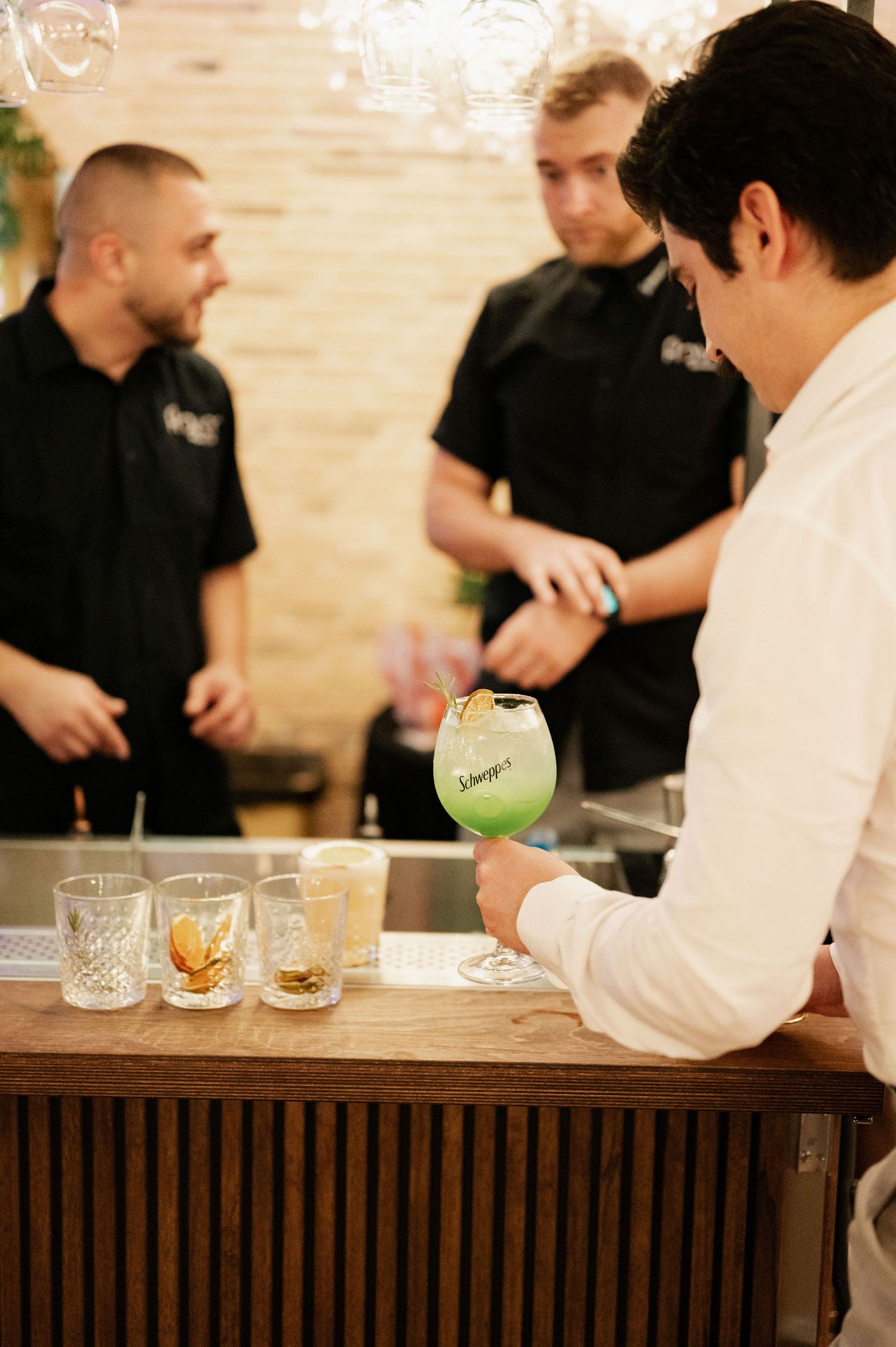 Bartenders in black uniforms prepare cocktails at a wooden bar counter while discussing drinks with customers.