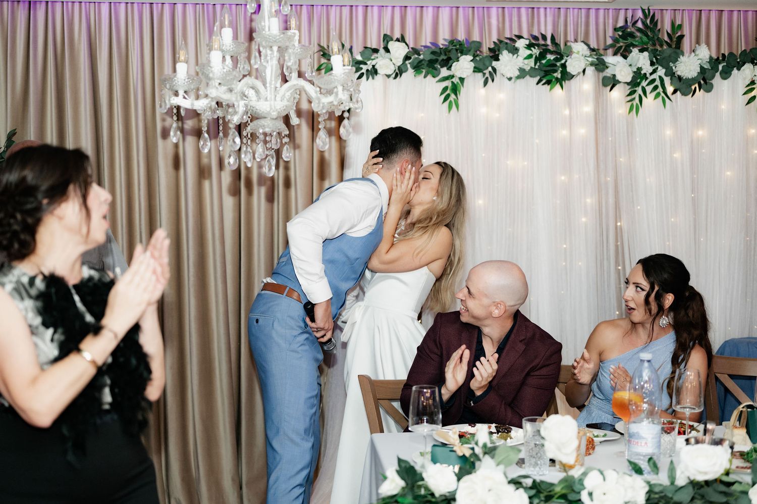 A romantic moment unfolds at a wedding reception table decorated with greenery and soft lighting.
