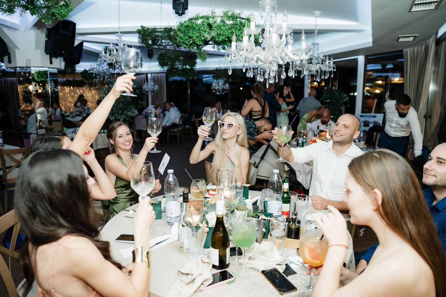 Wedding reception guests celebrate and dance together under elegant chandeliers at an indoor venue.