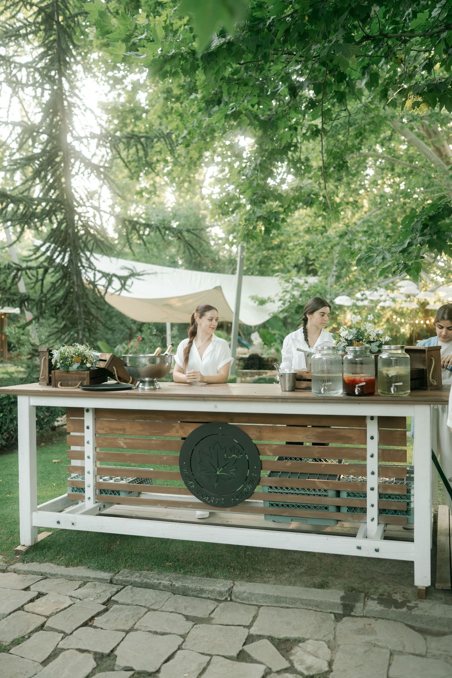 A rustic wooden bench with white metal supports sits under shady trees in an outdoor garden setting.