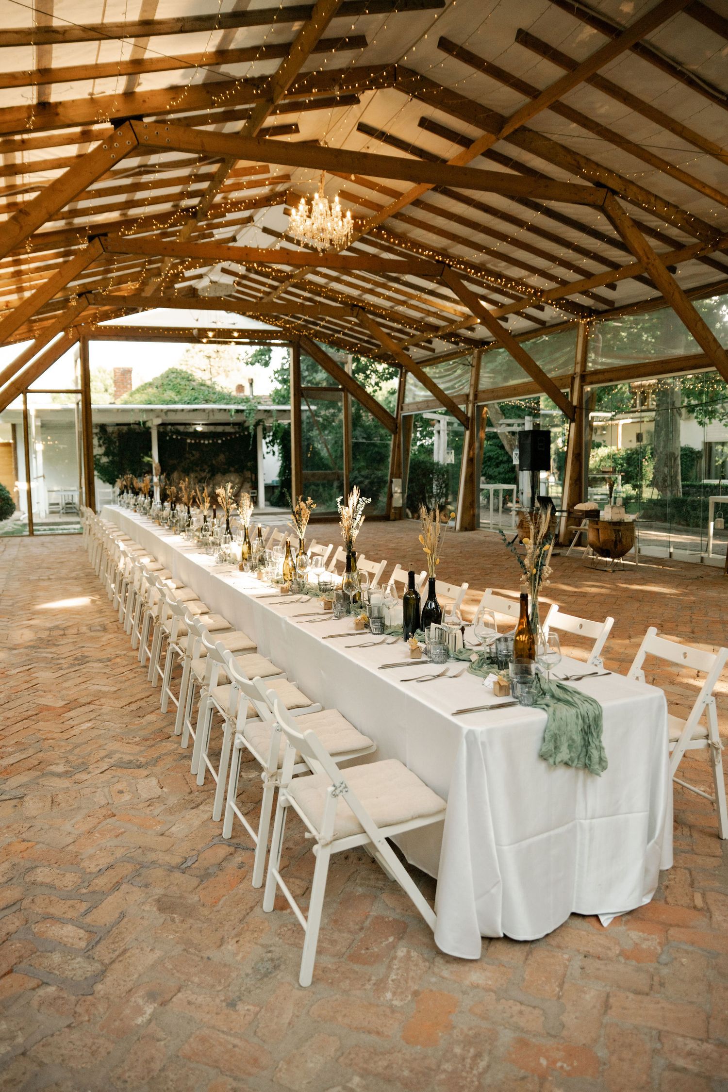 Long rustic covered venue with white chairs and tables set up for an elegant wedding reception on stone flooring.