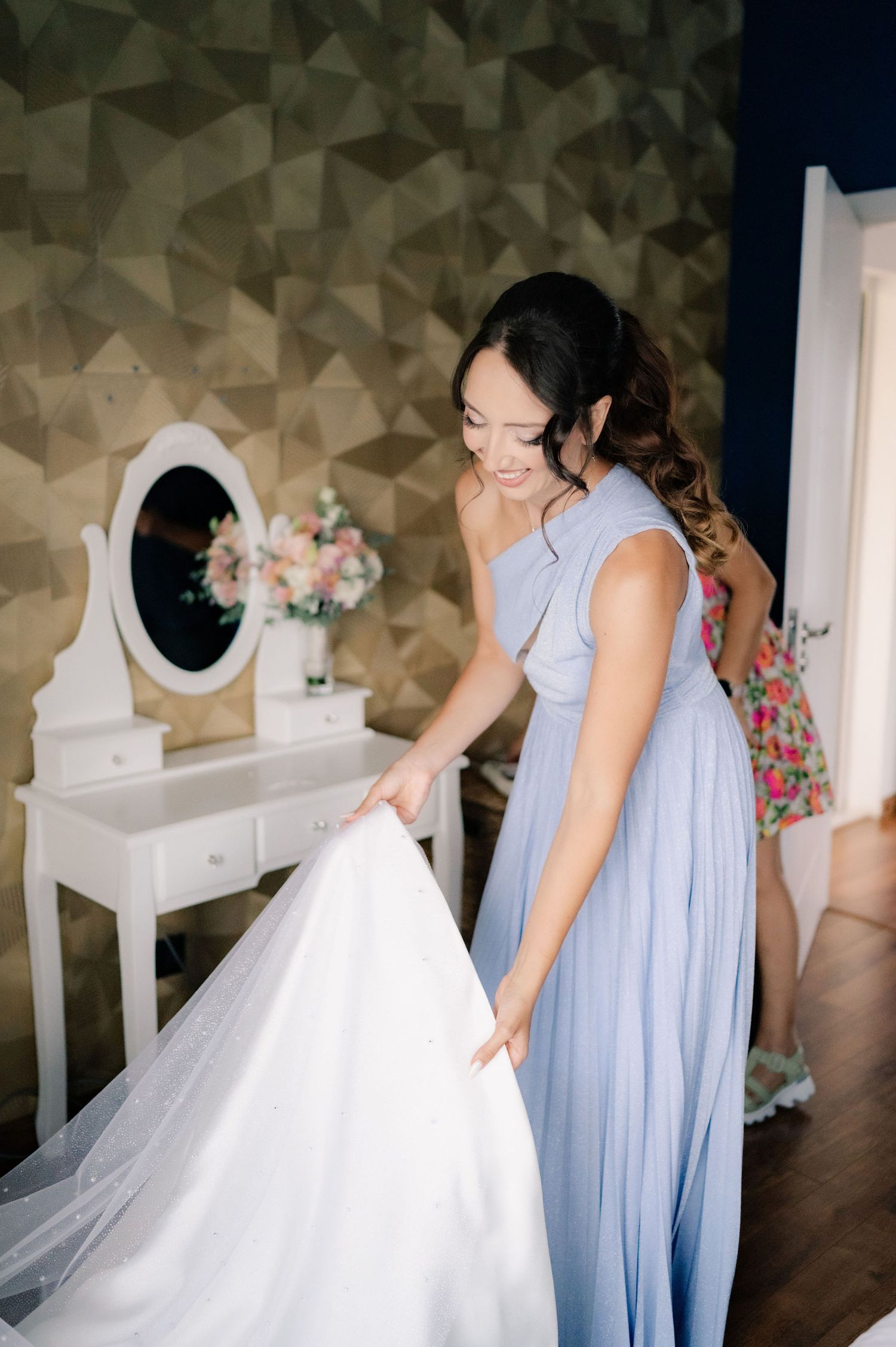 A person in a light blue bridesmaid dress helps adjust a white wedding gown train in a dressing room.