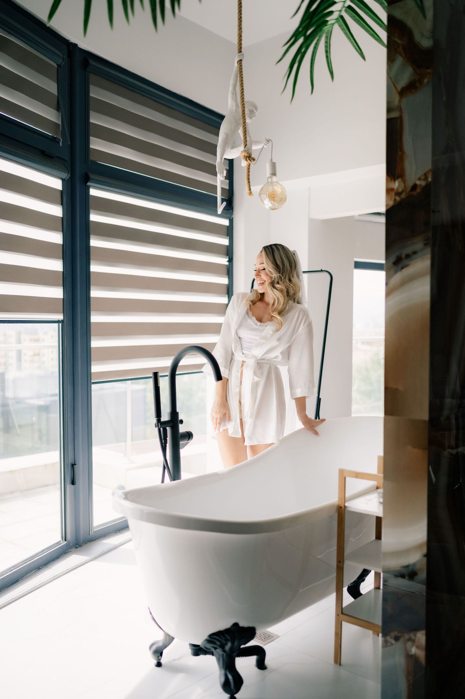 Modern bathroom interior with freestanding bathtub, black faucet, and natural light streaming through striped window shades.