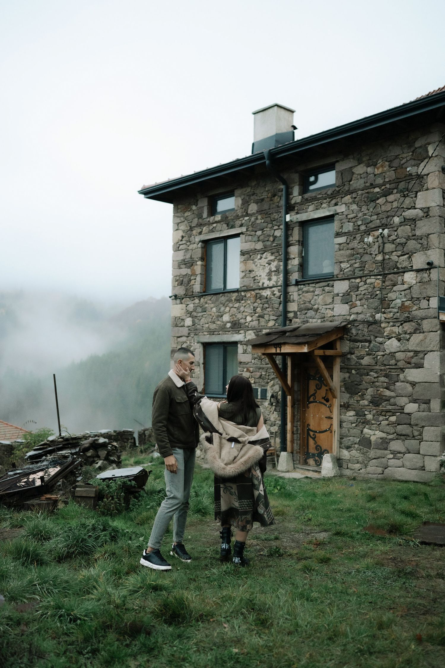Two people stand outside a rustic stone house with fog rolling in the background on a misty mountainside.