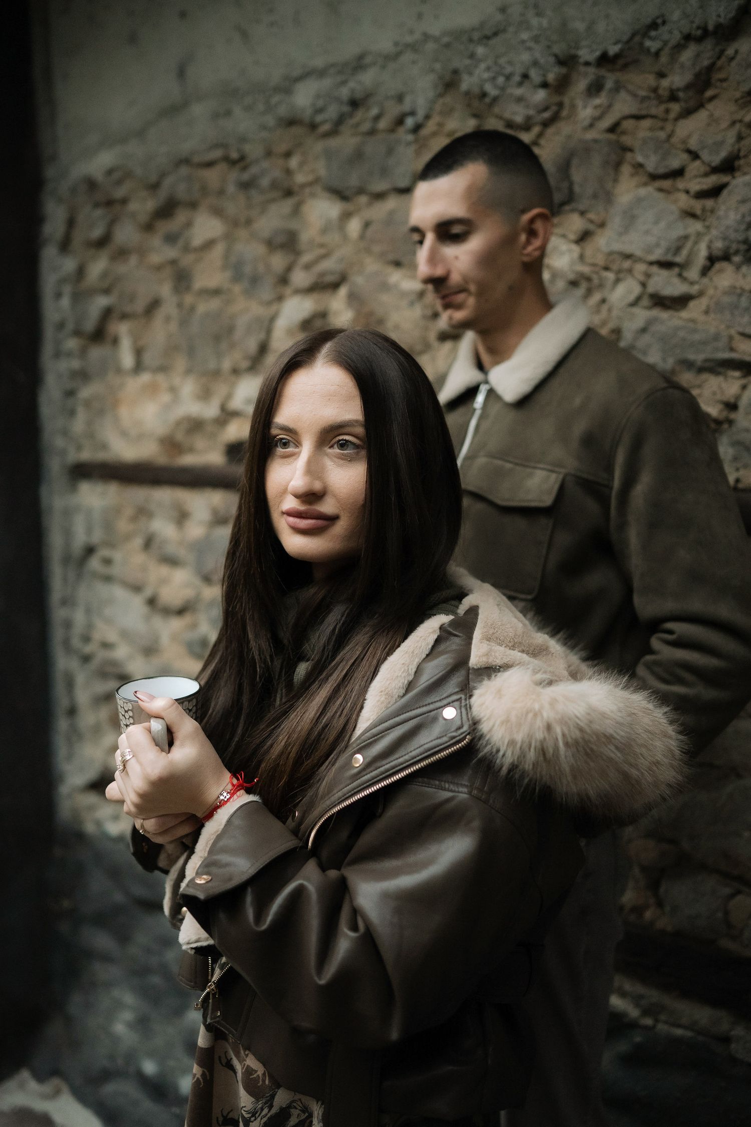 A couple in winter coats stands against a stone wall, with the woman holding a coffee cup in the foreground.