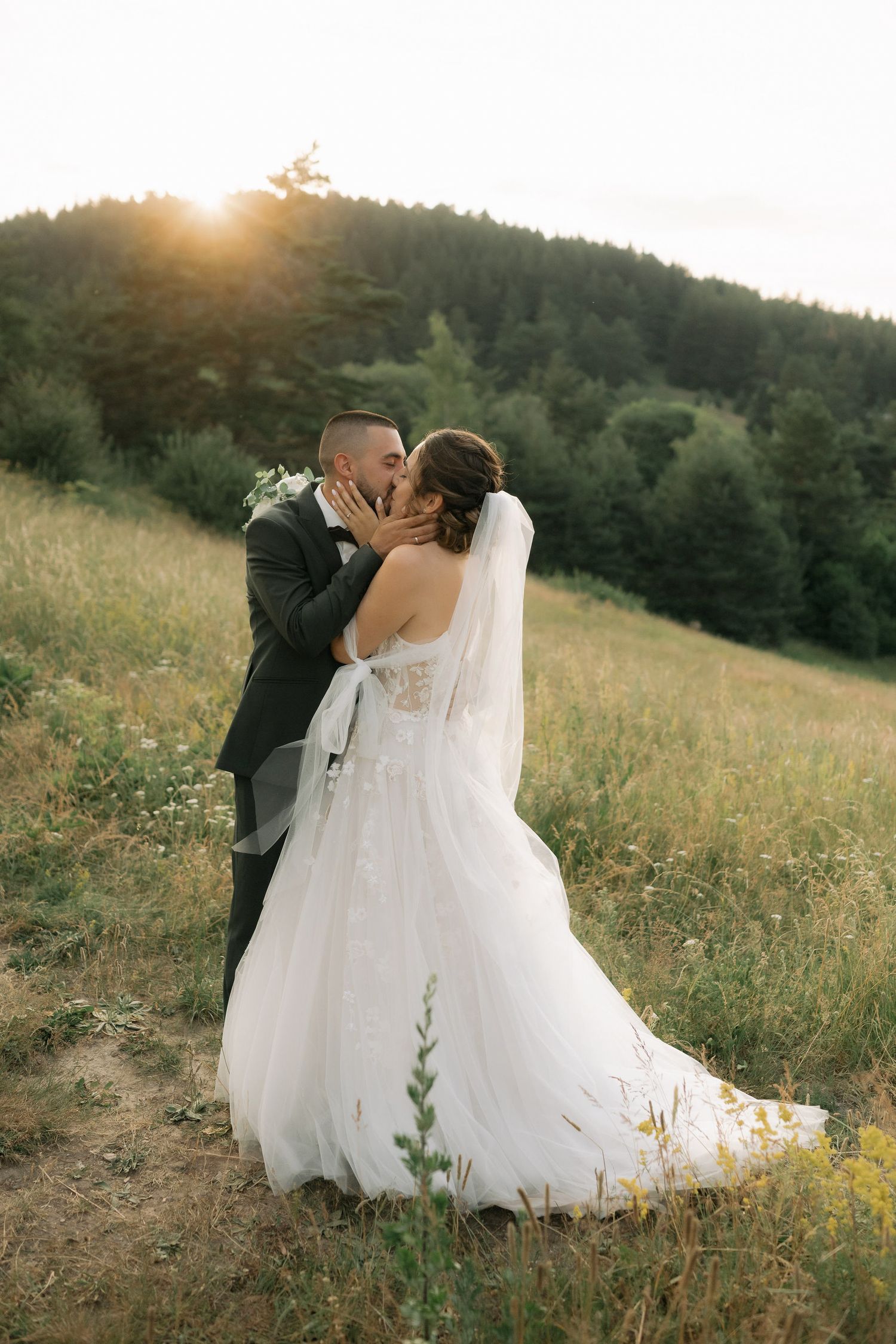 Romantic wedding couple embraces in a sunlit meadow at golden hour with rolling hills in the background.