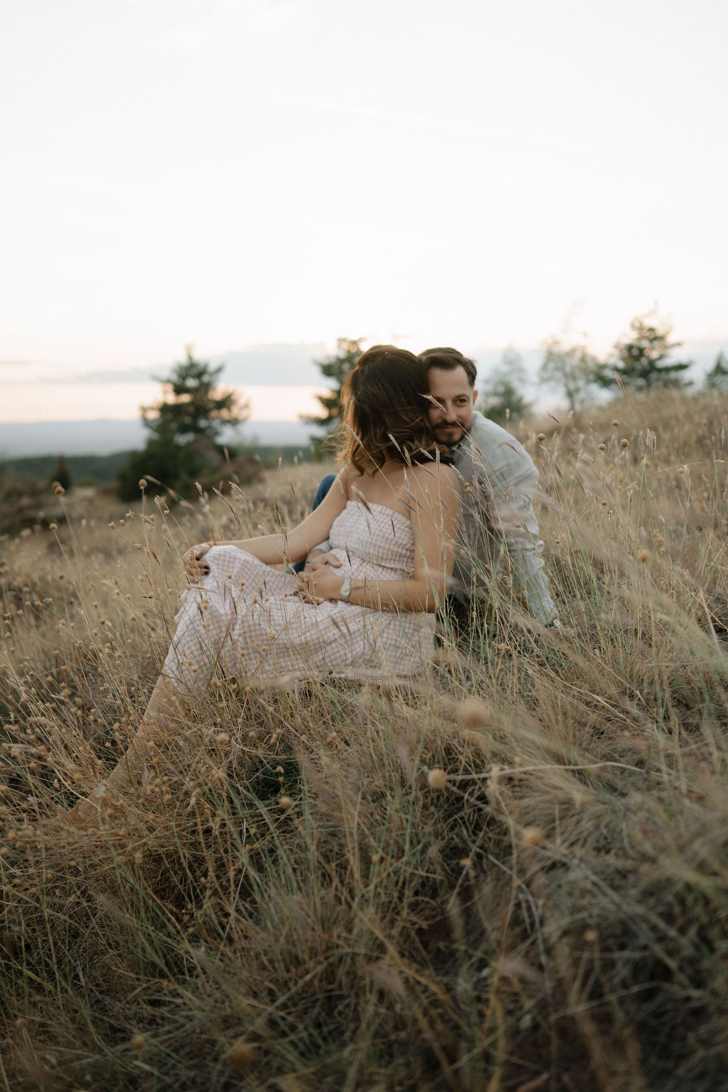 A couple embraces intimately while lying in tall grass during golden hour in a rustic outdoor setting.