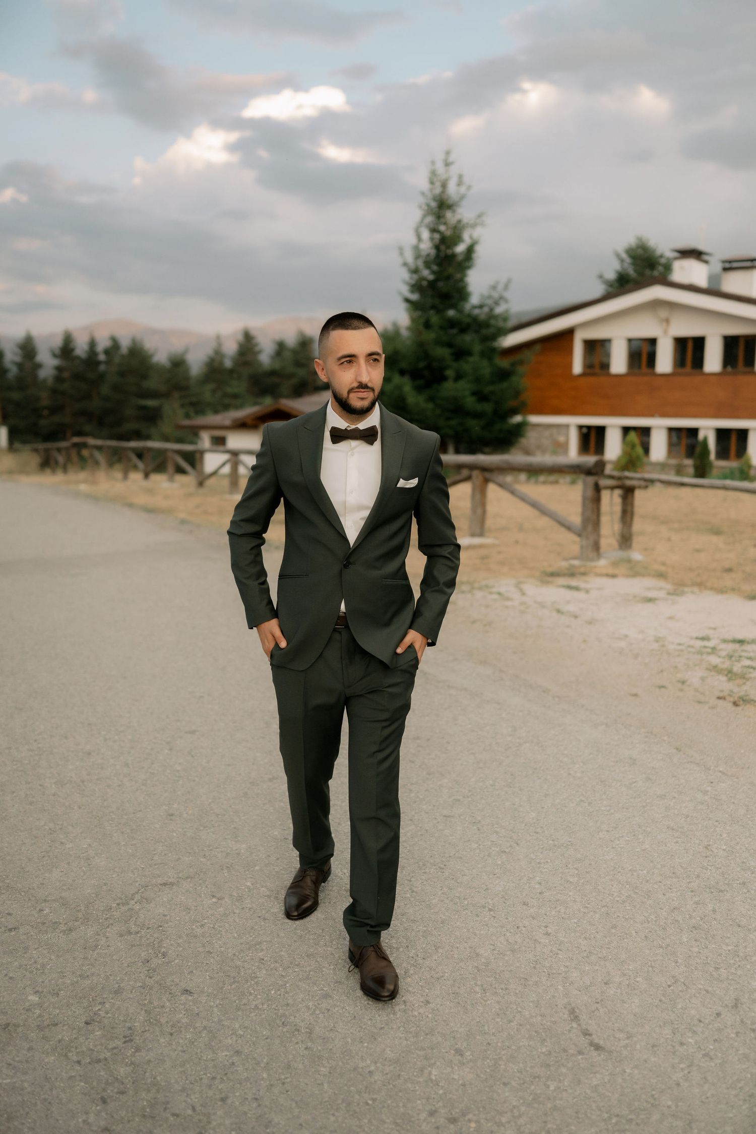 A person in a formal black tuxedo and bowtie walks along a gravel path on an overcast day.