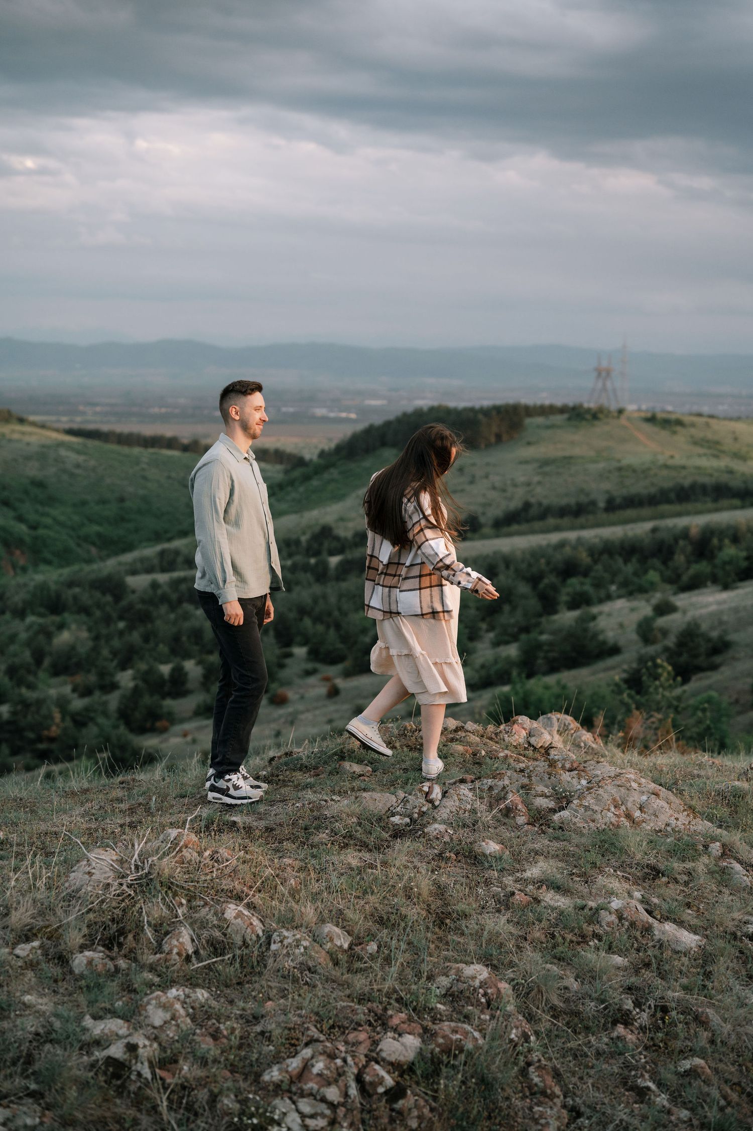 Figures traverse a rugged hilltop trail with expansive valley views beneath overcast skies.