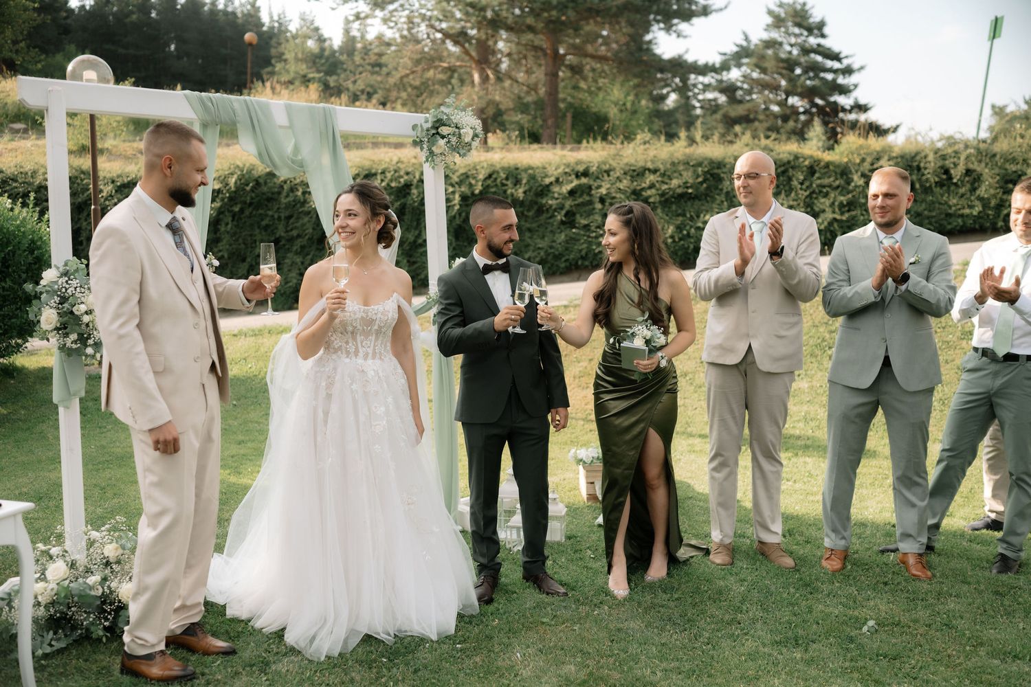 Wedding party in neutral tones stands together on grassy lawn celebrating during outdoor summer ceremony.