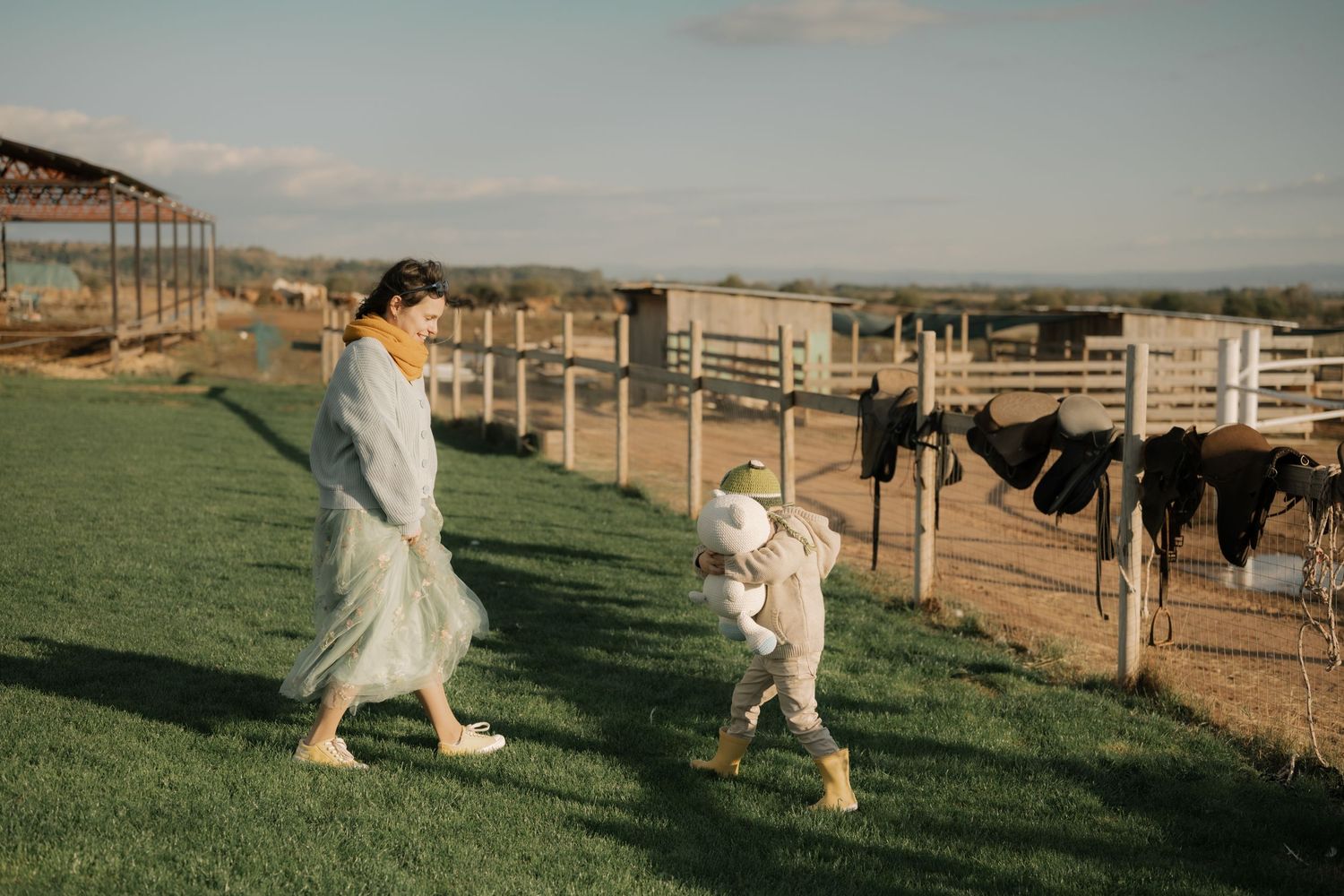 A couple in white wedding attire walks through a scenic countryside ranch with wooden fences and horses at sunset.