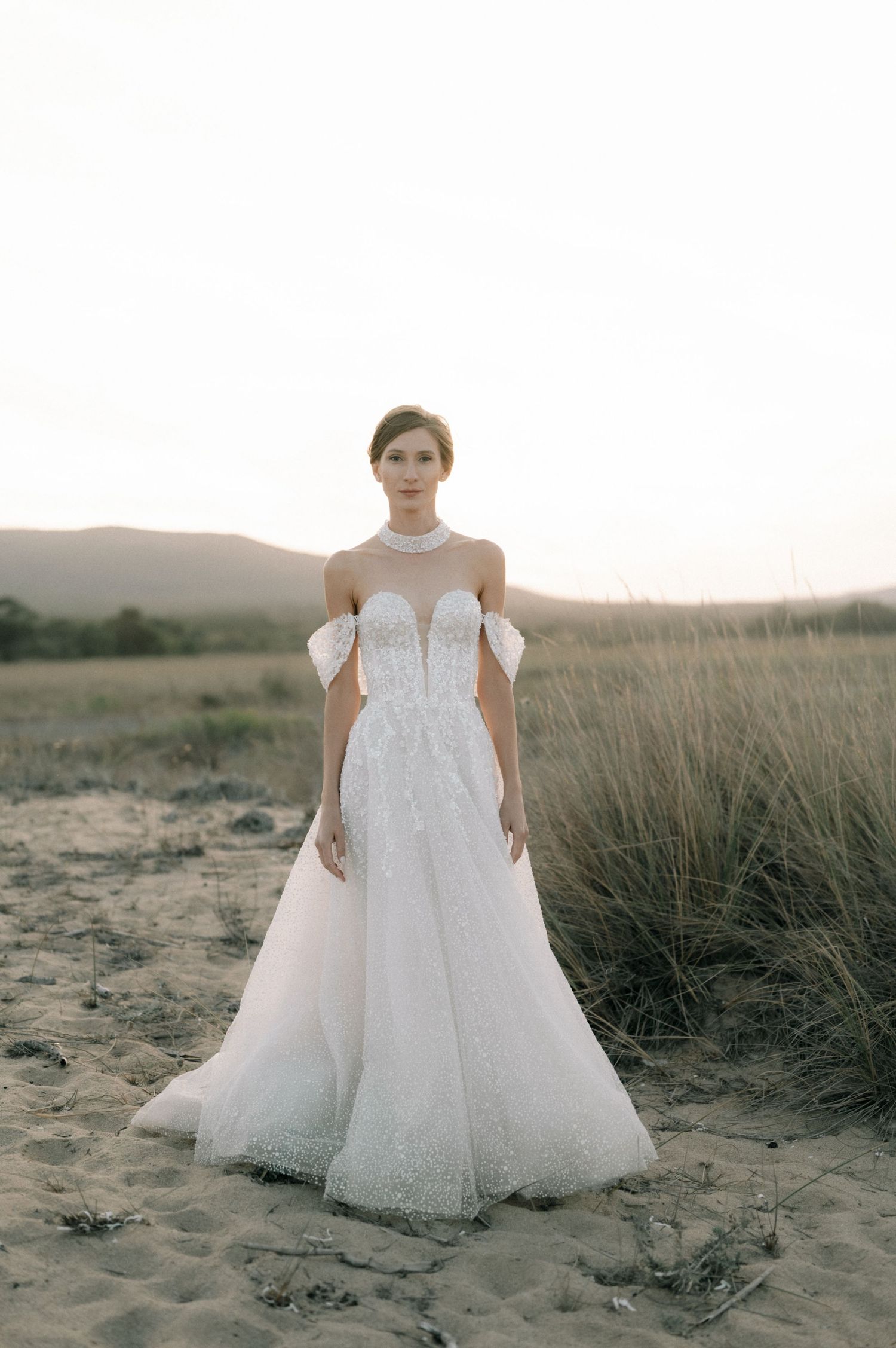 A bride in a stunning off-shoulder sparkly white A-line wedding gown poses gracefully on a sandy beach at dusk.