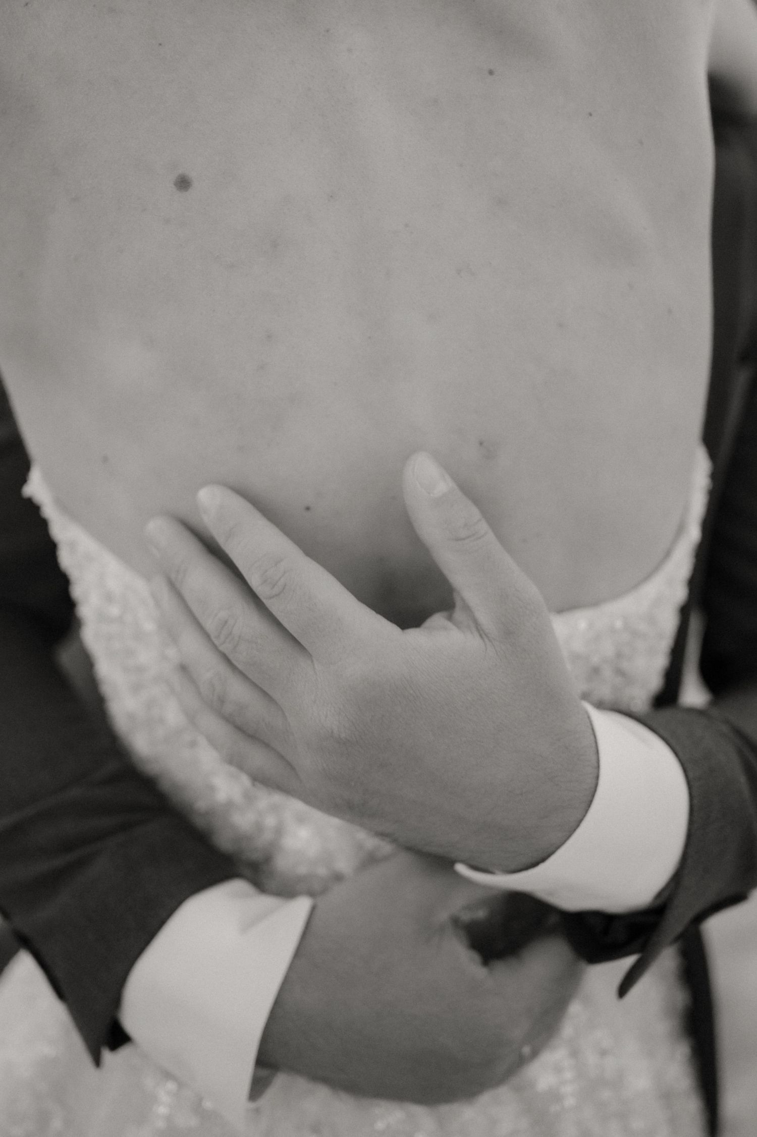Black and white photo of a groom's hand resting on a bride's bare back in a lace wedding dress.