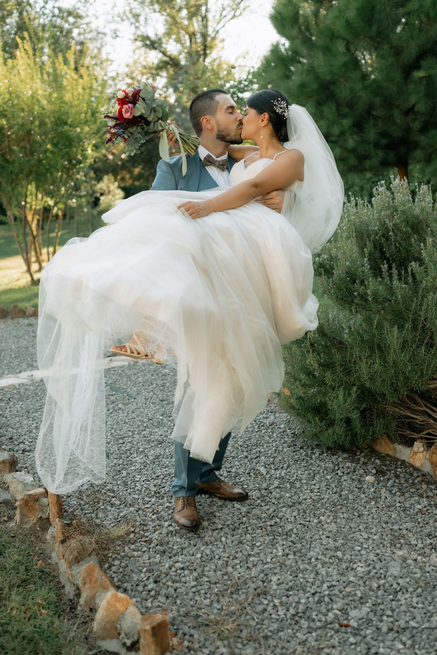 A romantic wedding couple shares a dip kiss on a gravel path while their small dog watches in a garden setting.