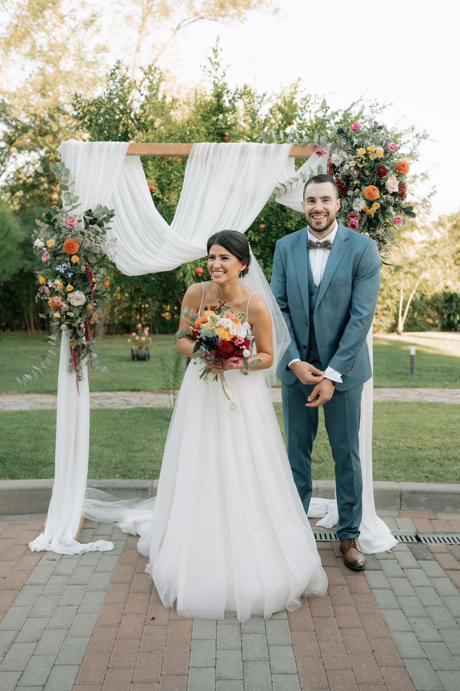 A couple stands under a floral wedding arch with white draping and colorful flowers during an outdoor ceremony.