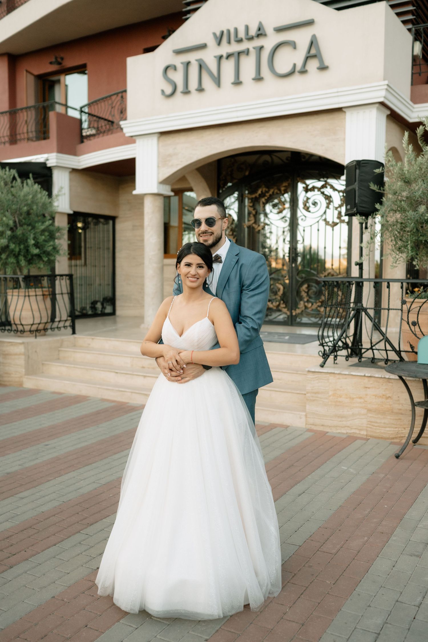 A wedding couple poses in front of Villa Sintica entrance with brick pavers and wrought iron details.