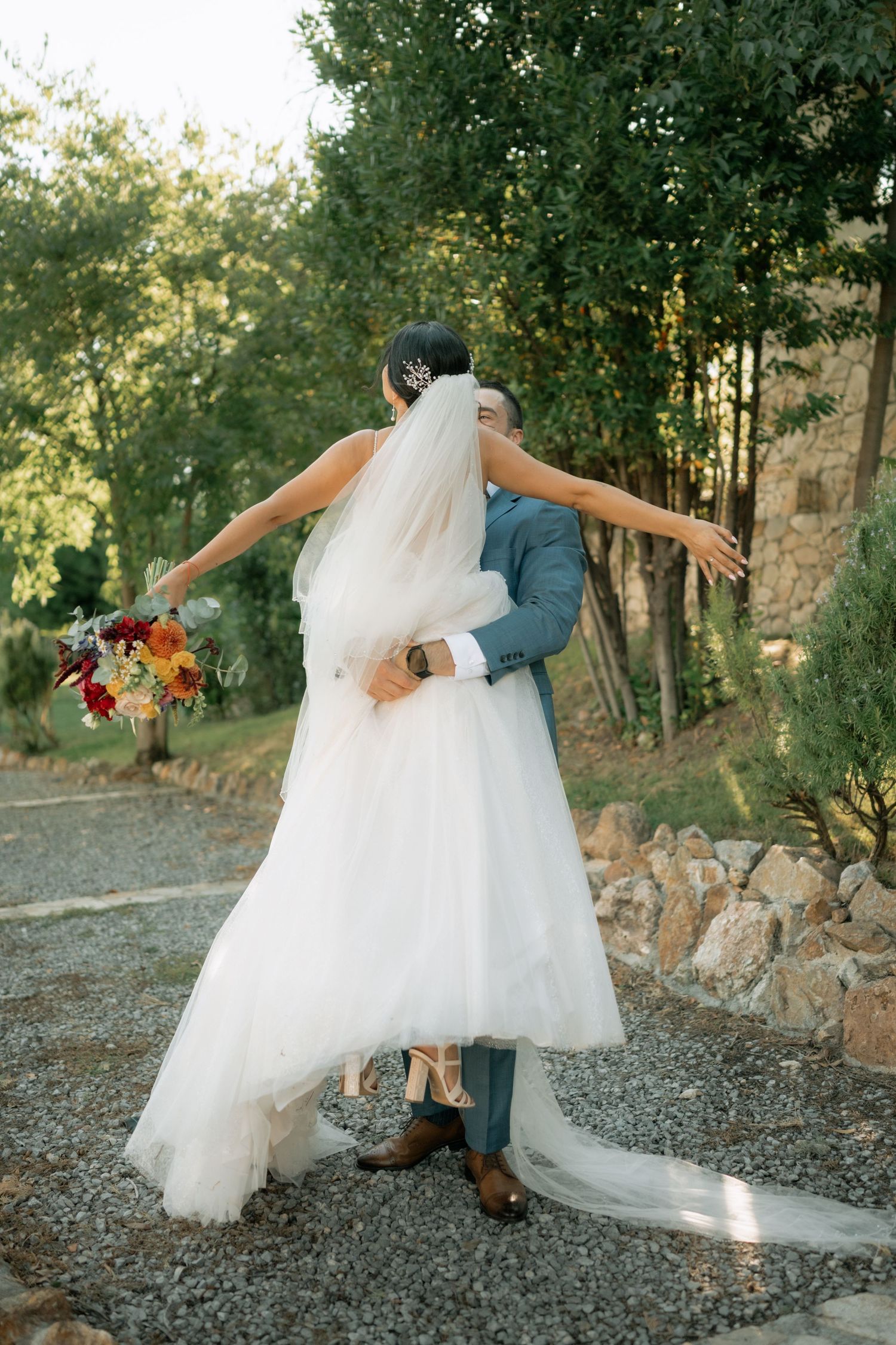 A romantic scene unfolds as a couple in wedding attire twirls playfully on a gravel path beside stone walls.