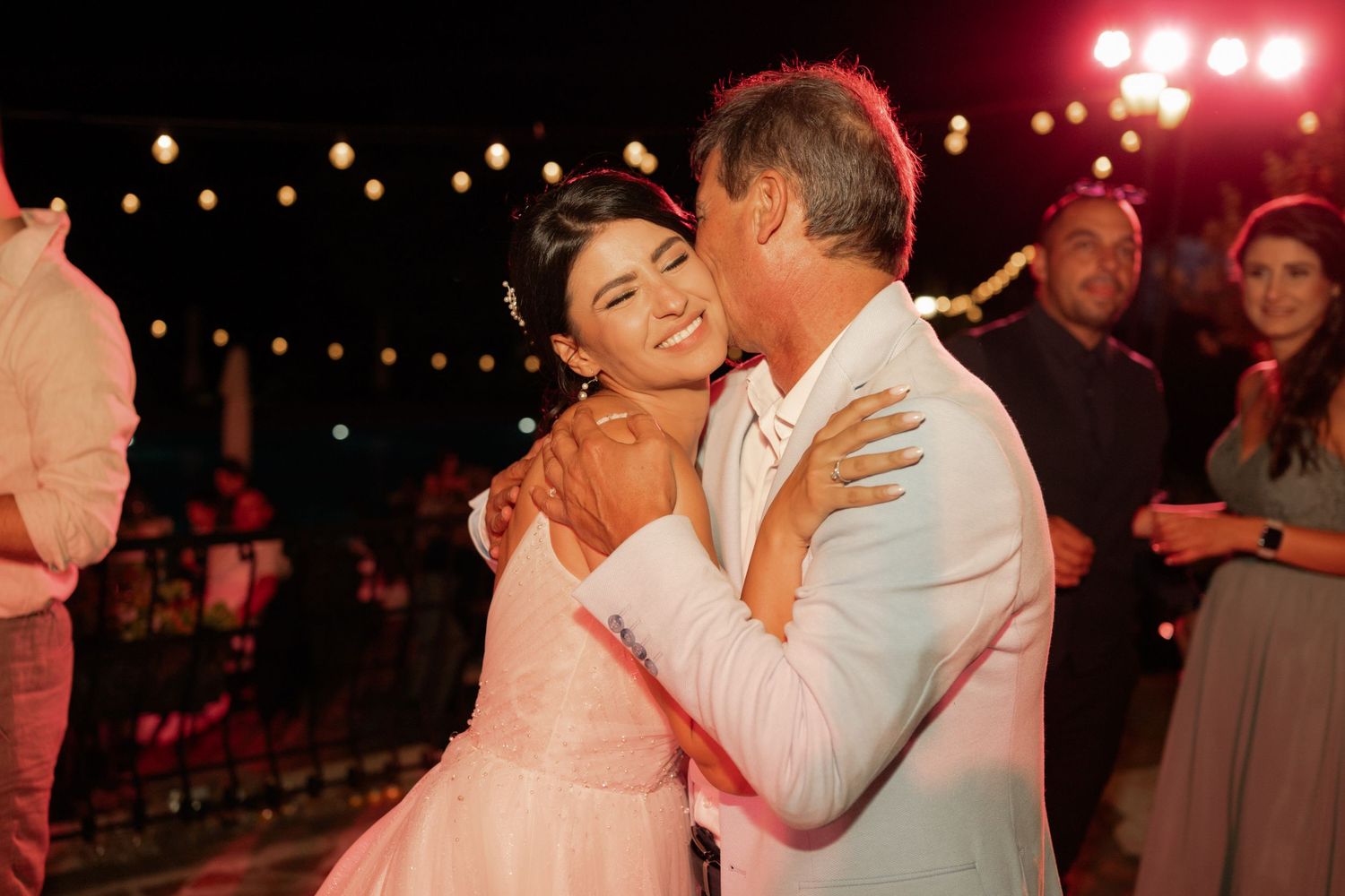 A couple shares a romantic dance moment under warm string lights at an evening outdoor reception.