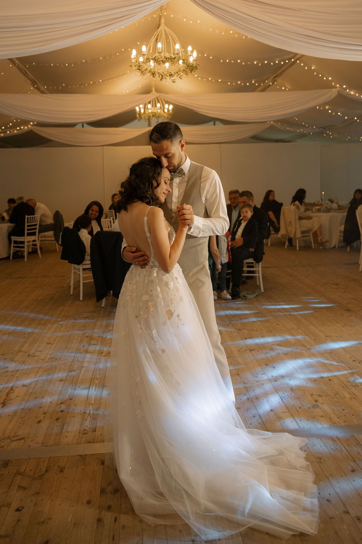 Bride and groom share their first dance under a chandelier in an elegant tent reception venue with fairy lights.