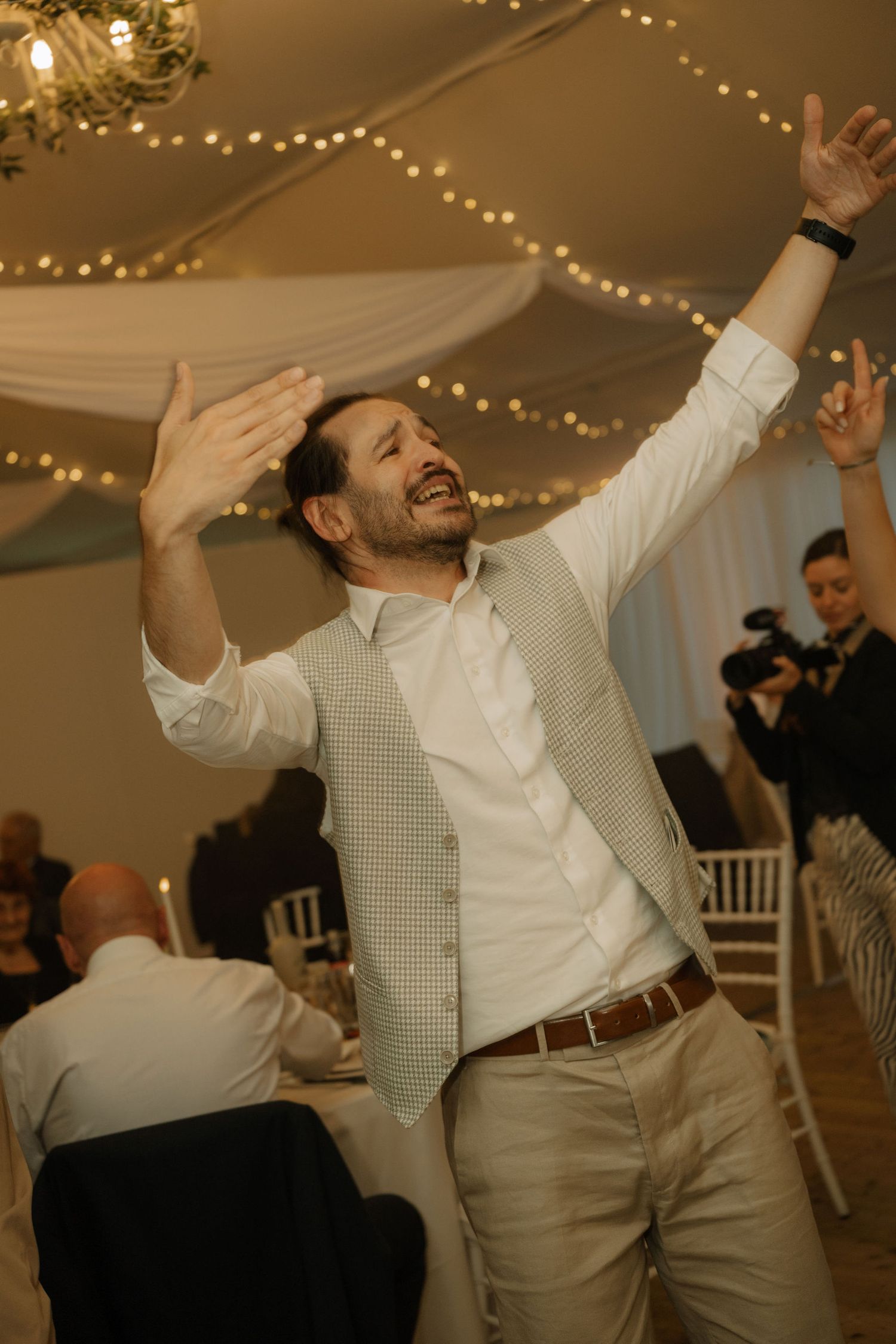 A joyful man dancing with arms raised at an elegant wedding reception venue with fairy lights.
