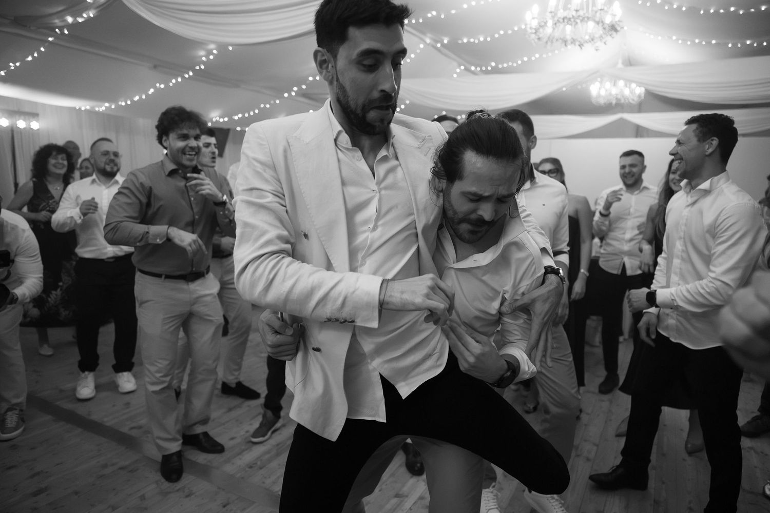 Black and white photo of two men dancing energetically at a wedding reception with guests in background.