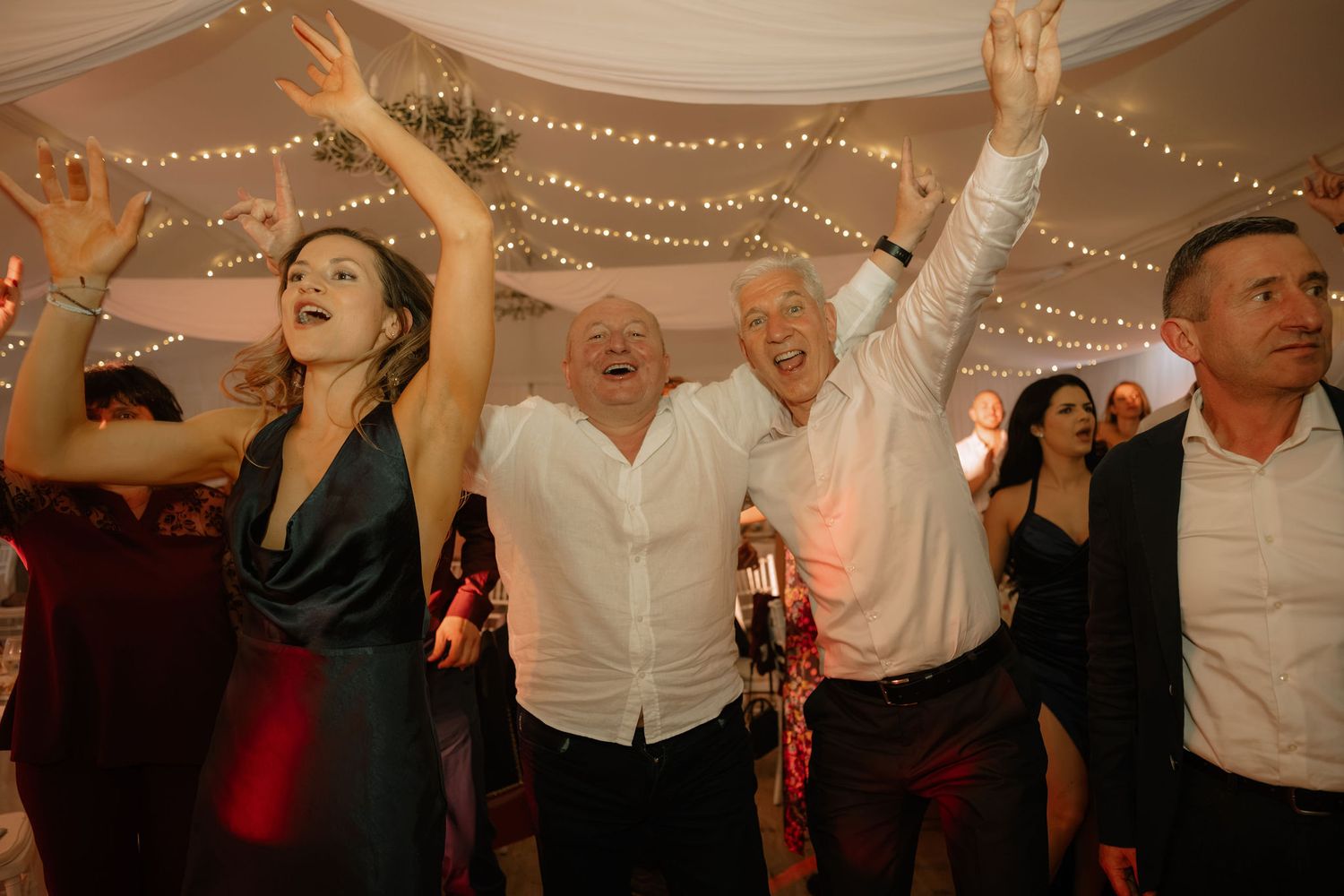 Guests dancing joyfully at a wedding reception under warm string lights in a decorated tent venue.