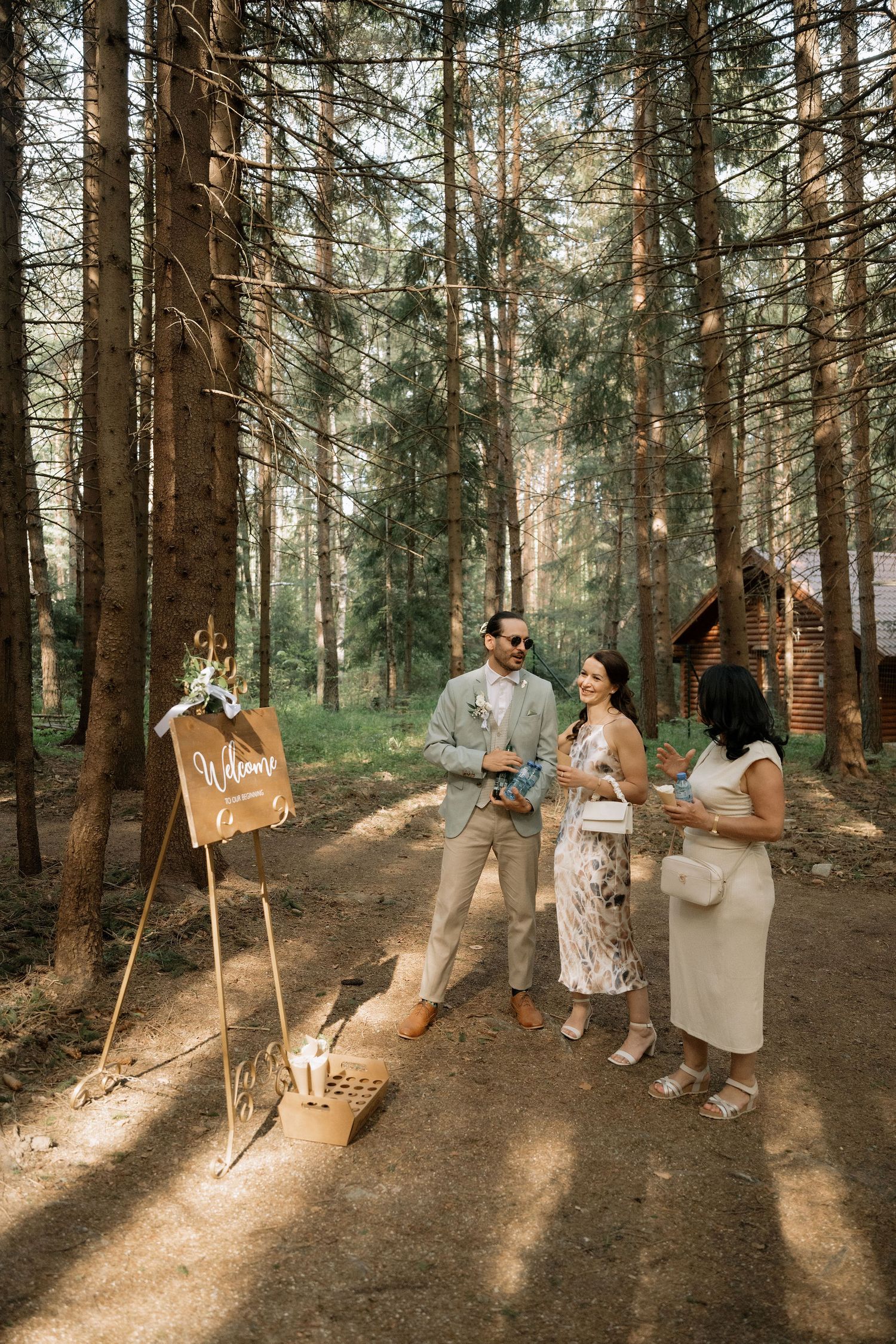 Outdoor wedding welcome sign on easel with couple and guest chatting in sunlit pine forest near rustic cabin.
