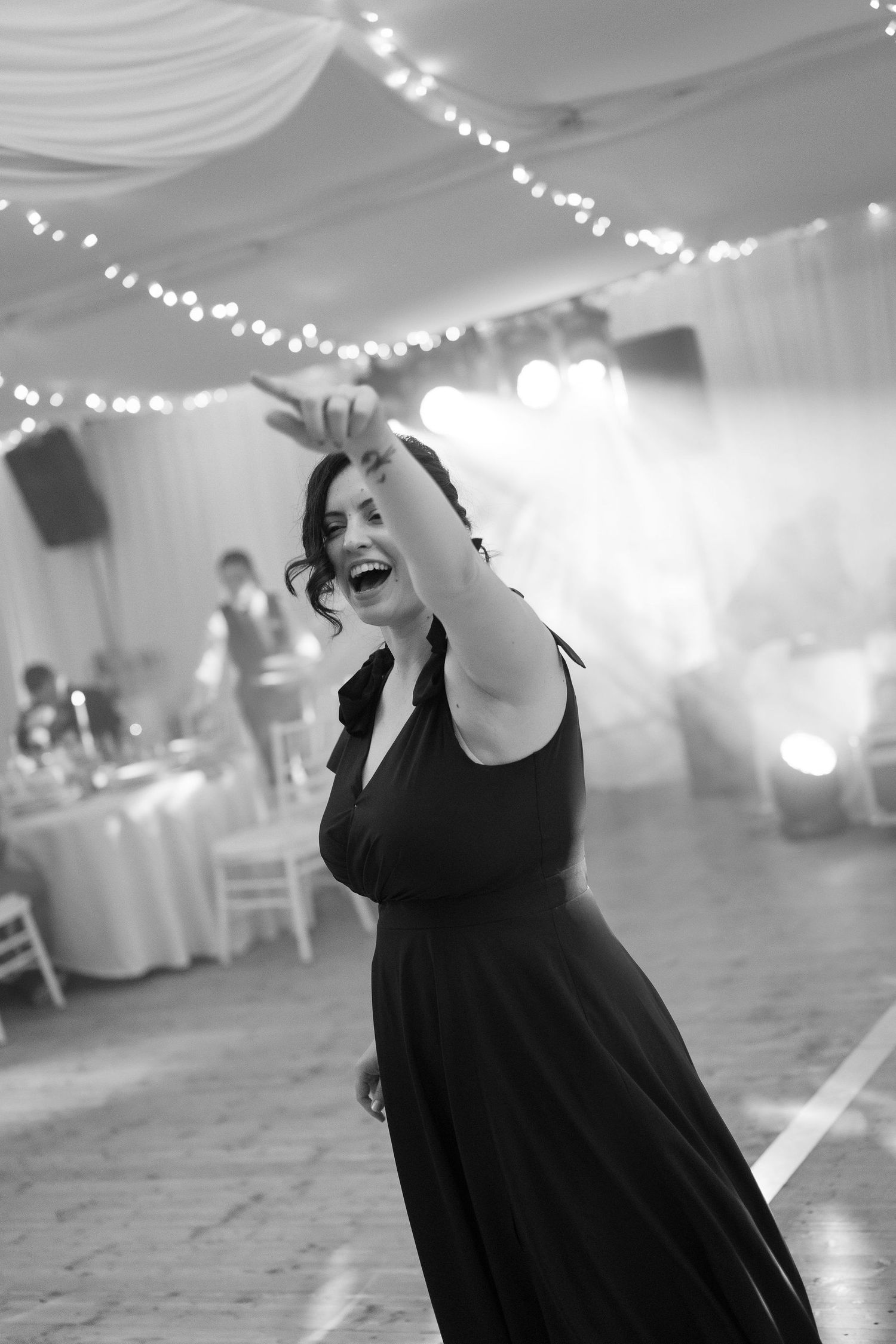 Woman in black dress dancing joyfully at a wedding reception with string lights in black and white photo.