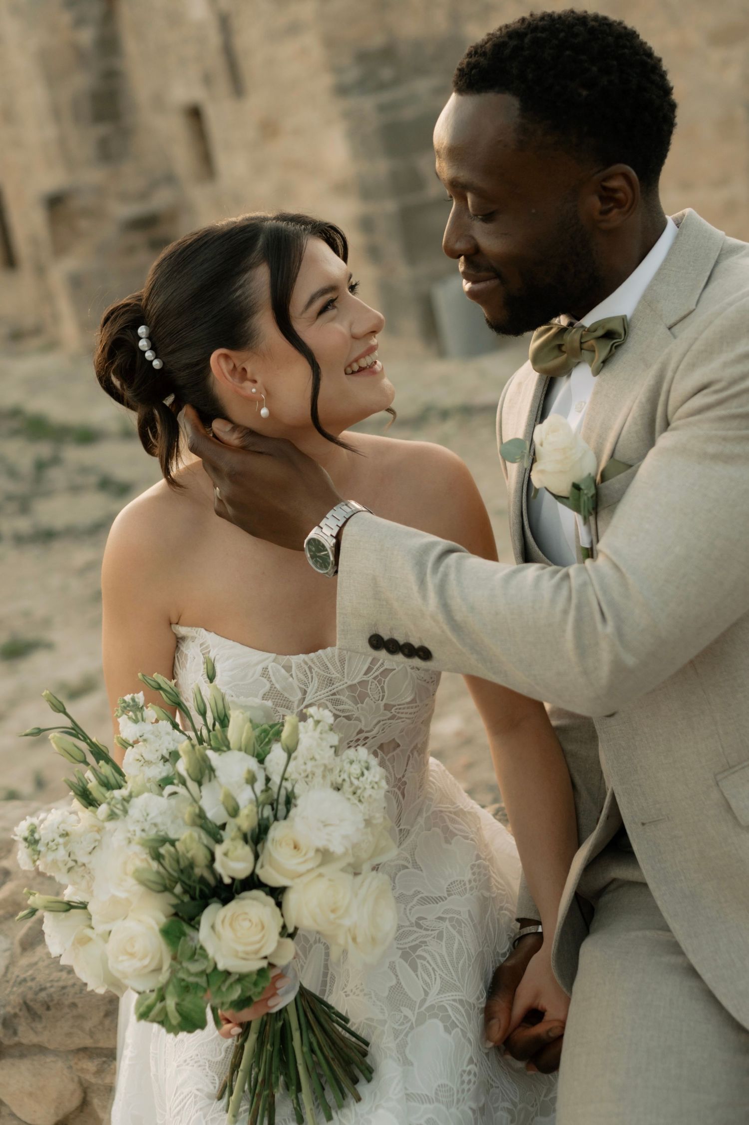 A groom in a beige suit tenderly cups his bride's face as she smiles, holding a white floral bouquet at an outdoor wedding.